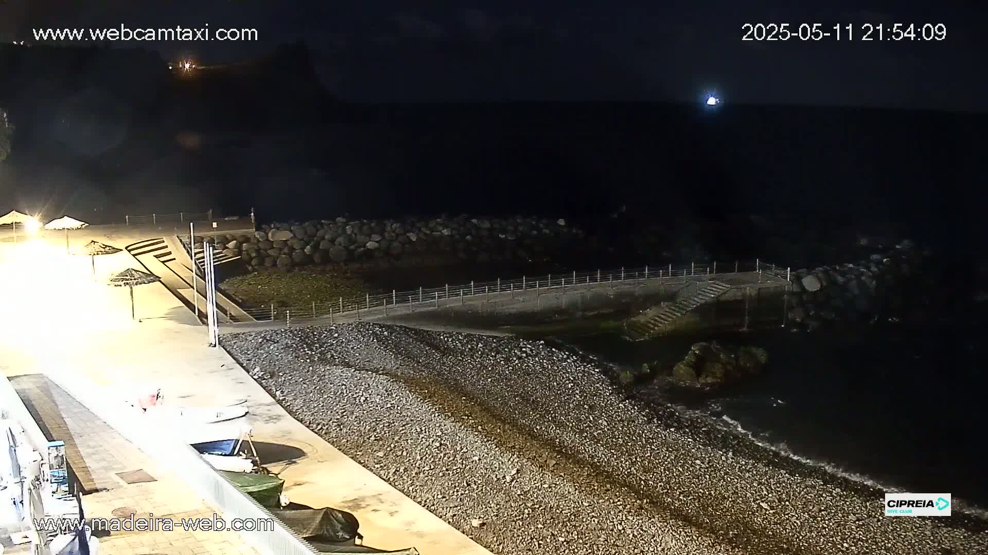 A dark, nighttime beach scene shows a pebble shoreline, a low wall, and calm, dark water.