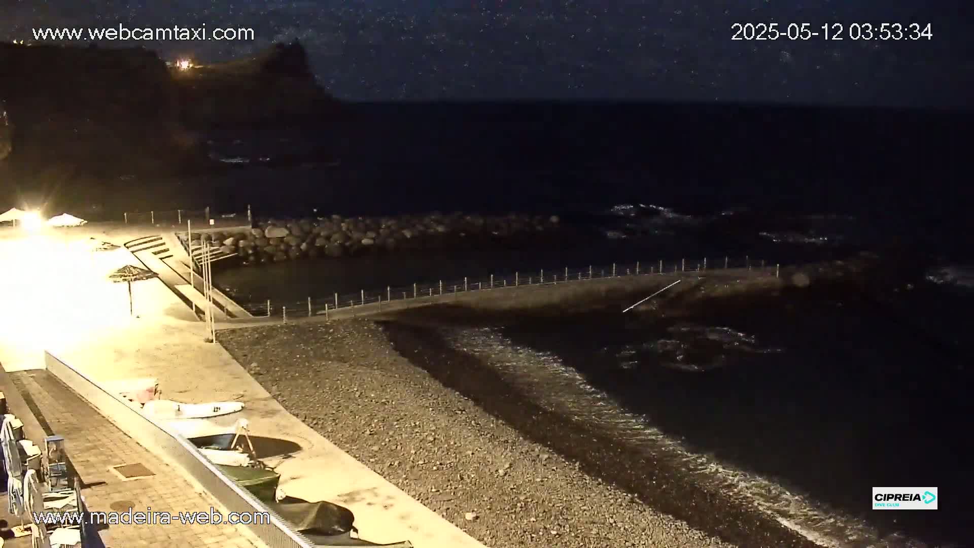 A nighttime view of a dark pebble beach with calm waves lapping the shore, a small harbor, and some boats are moored nearby under a slightly starlit sky.