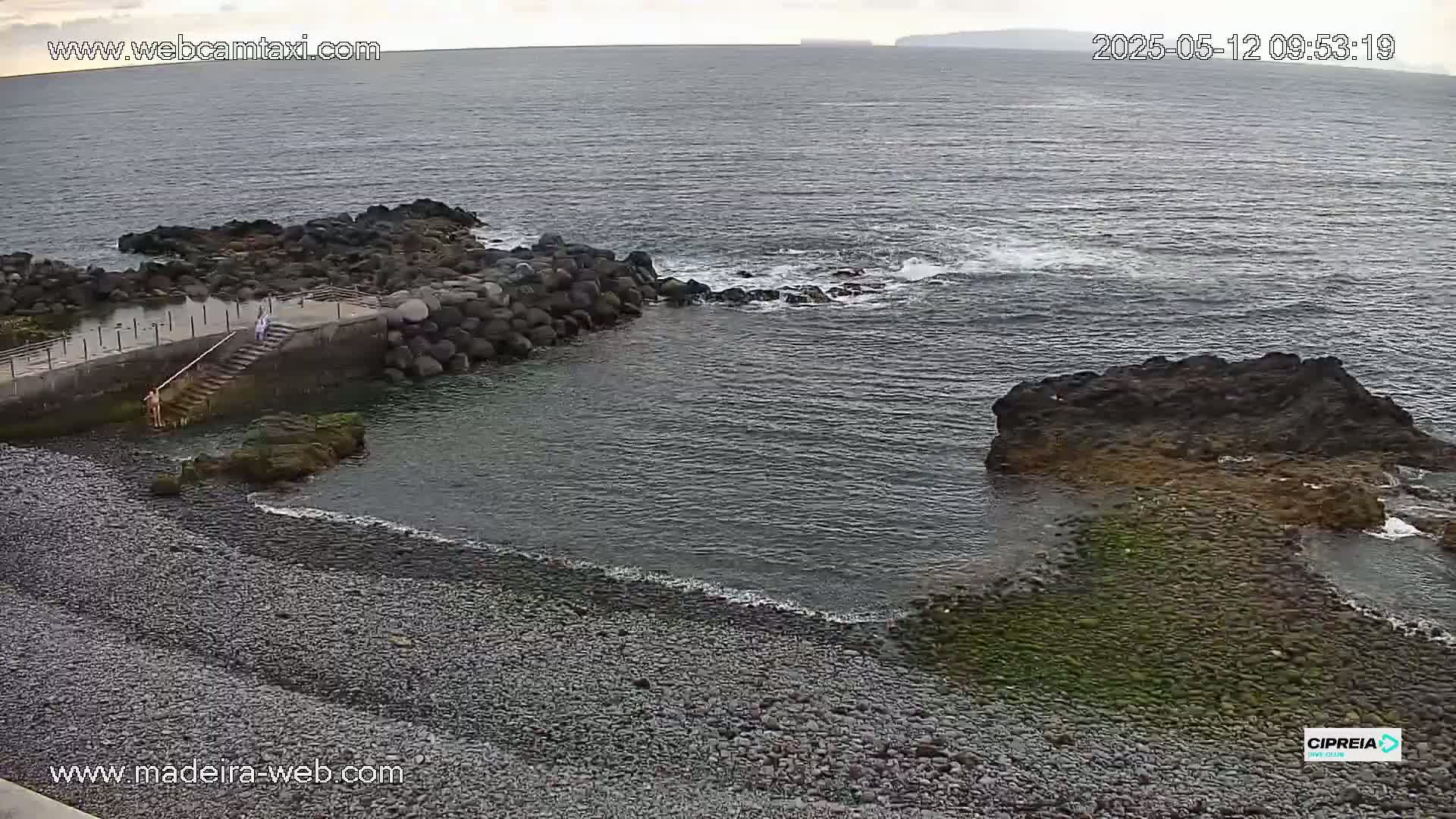 A rocky, pebble beach meets a calm ocean with a small stone jetty and a few people near the water's edge under an overcast sky.