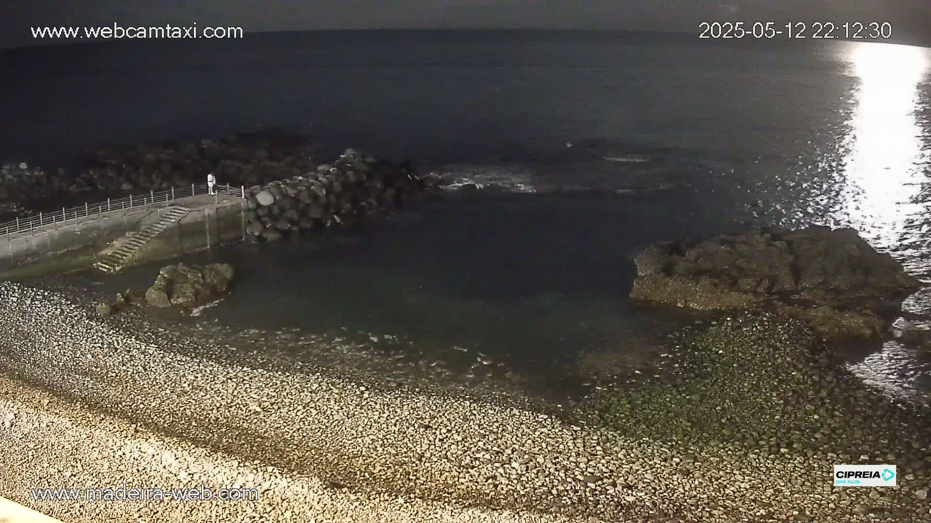 A rocky shoreline at night, partially lit by moonlight reflecting on the dark water, shows a small stone jetty with steps leading down to the water.
