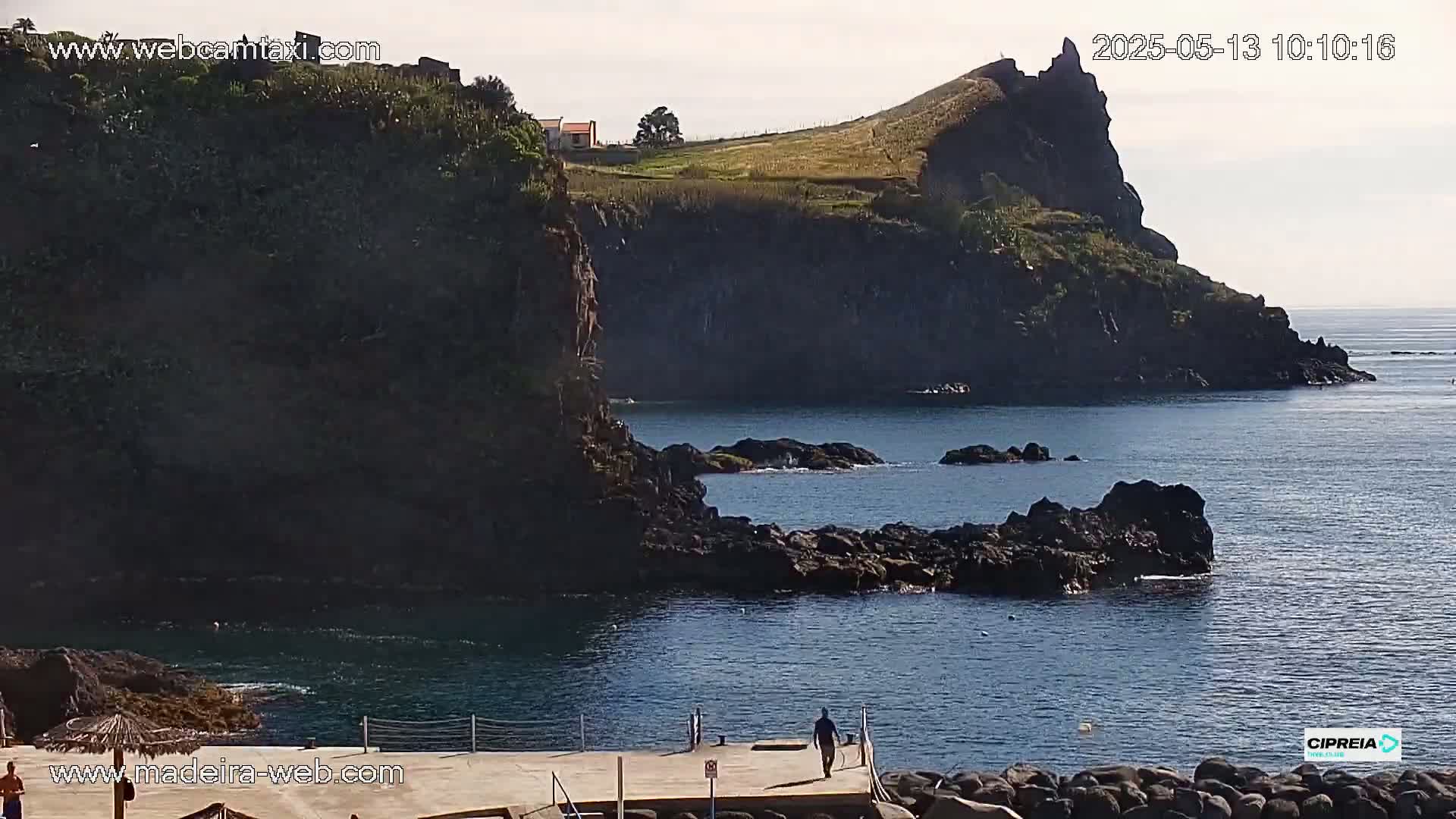 A person walks on a dock overlooking a calm ocean next to dark rocky cliffs and a small house on a grassy hill under a partly cloudy sky.