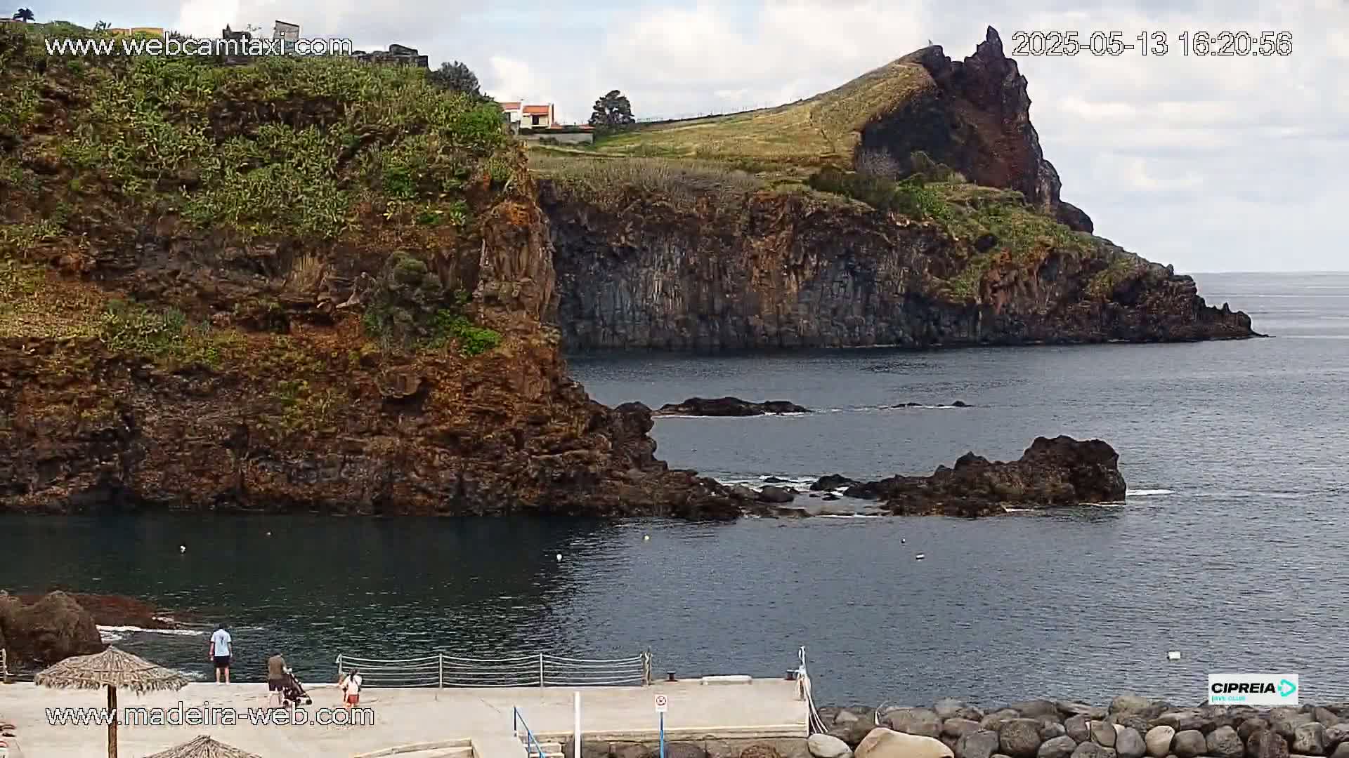 A rocky, dark-colored coastline with a small, calm bay, featuring a few people on a cement dock under a partly cloudy sky.