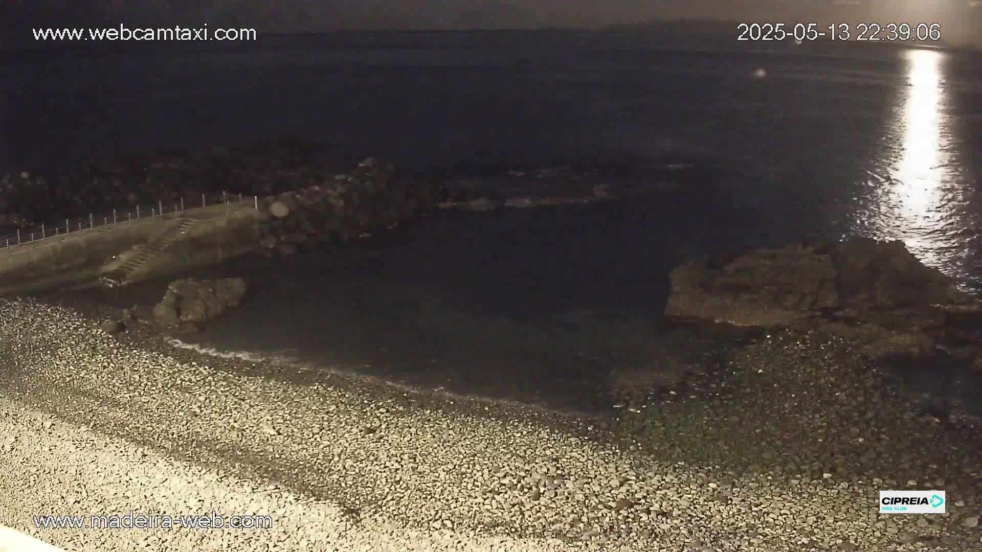 A rocky shoreline at night, under a bright moon, shows a pebble beach meeting dark water.