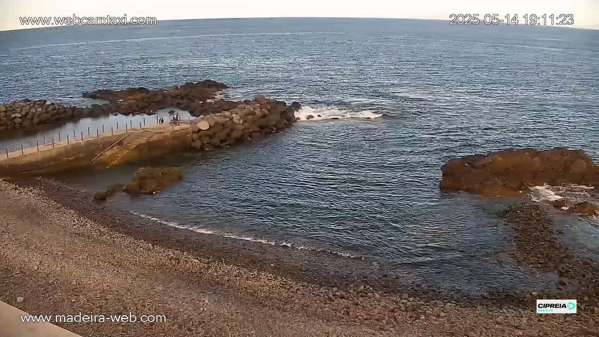 A rocky shoreline with calm water, a small stone jetty with a few people on it, and a pebble beach.