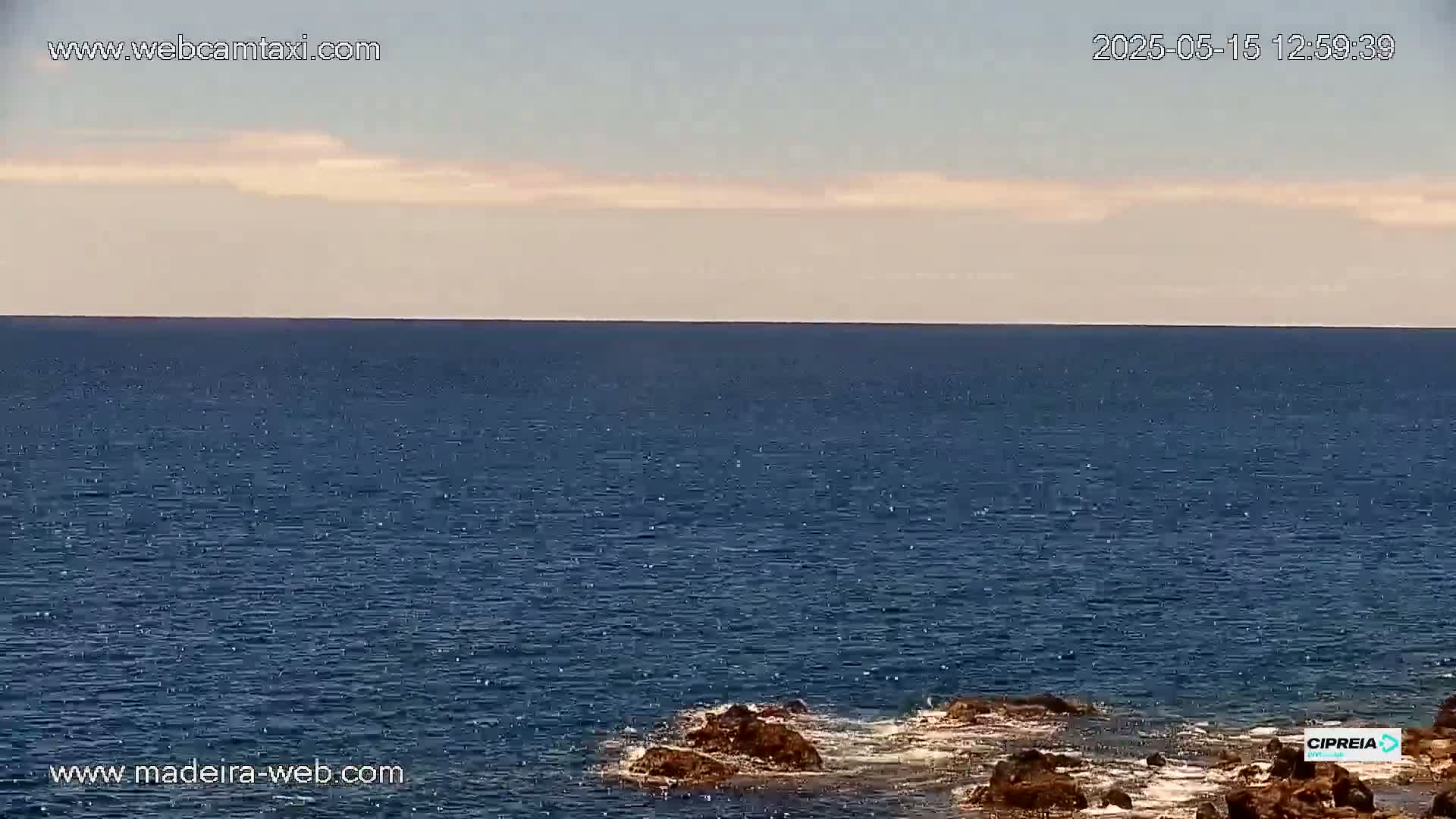 A mostly calm, dark blue ocean stretches to a pale, clear sky under mostly sunny conditions with a few wispy clouds, meeting rocky shores in the foreground.