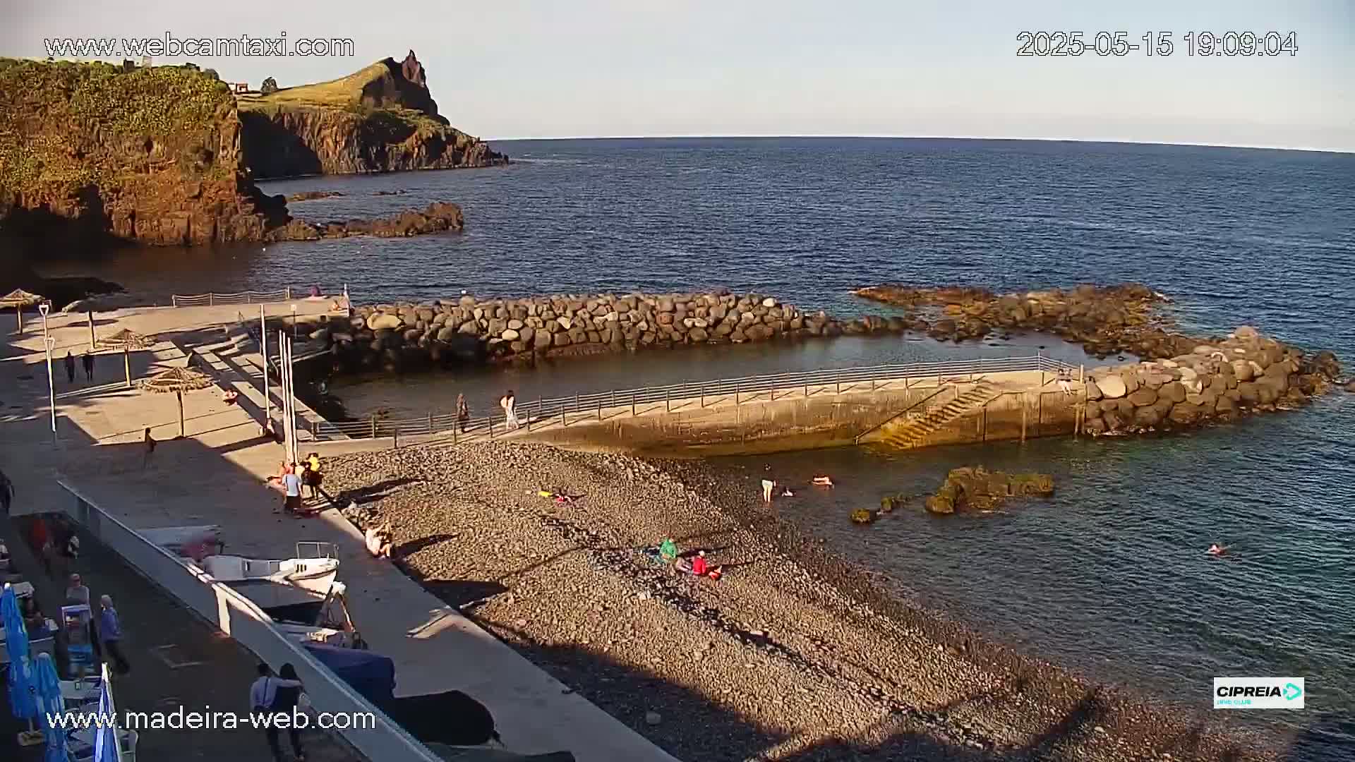 A rocky coast with a small, calm cove, a pebble beach, and several people relaxing and swimming under a clear, sunny sky.