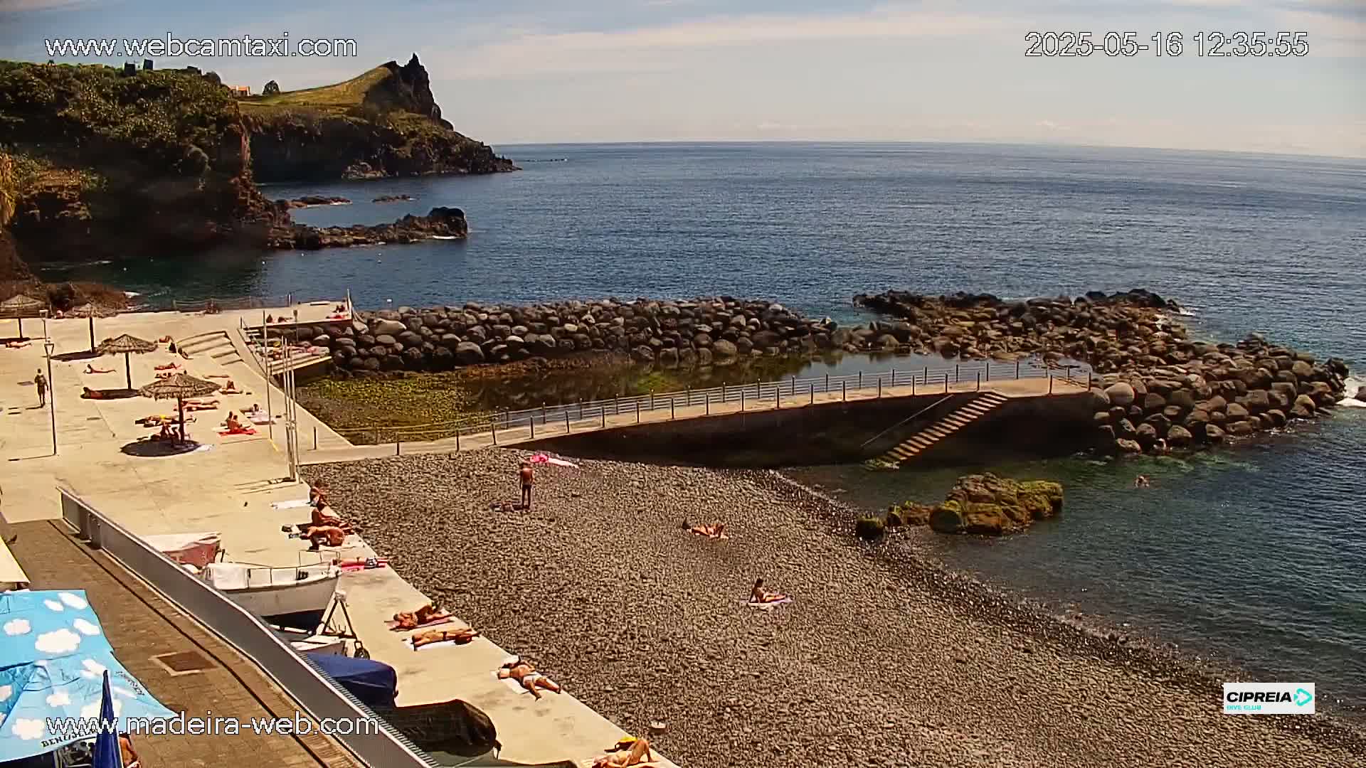 A rocky beach with sunbathers, a small man-made pool, and a calm ocean under a mostly sunny sky.