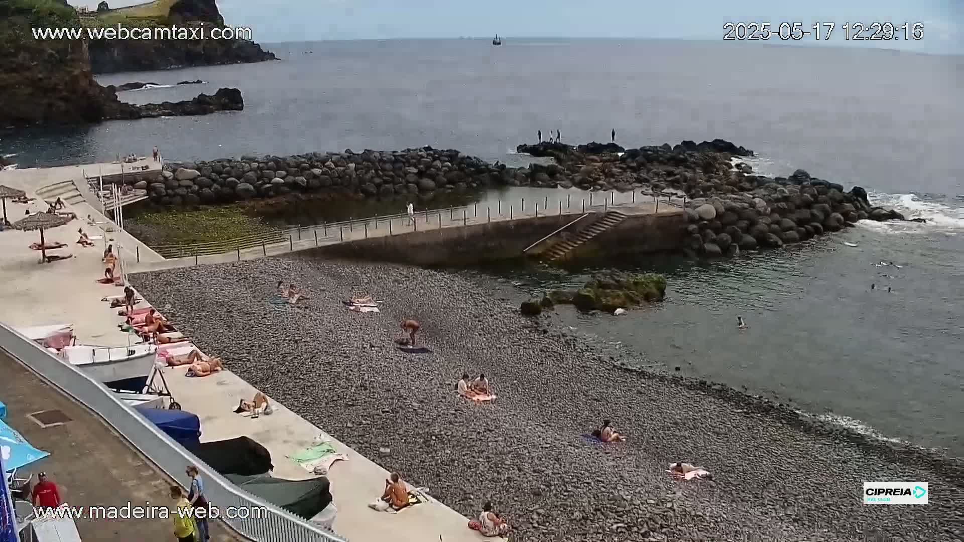 A pebble beach with a few people sunbathing and swimming, next to a calm ocean on an overcast day.