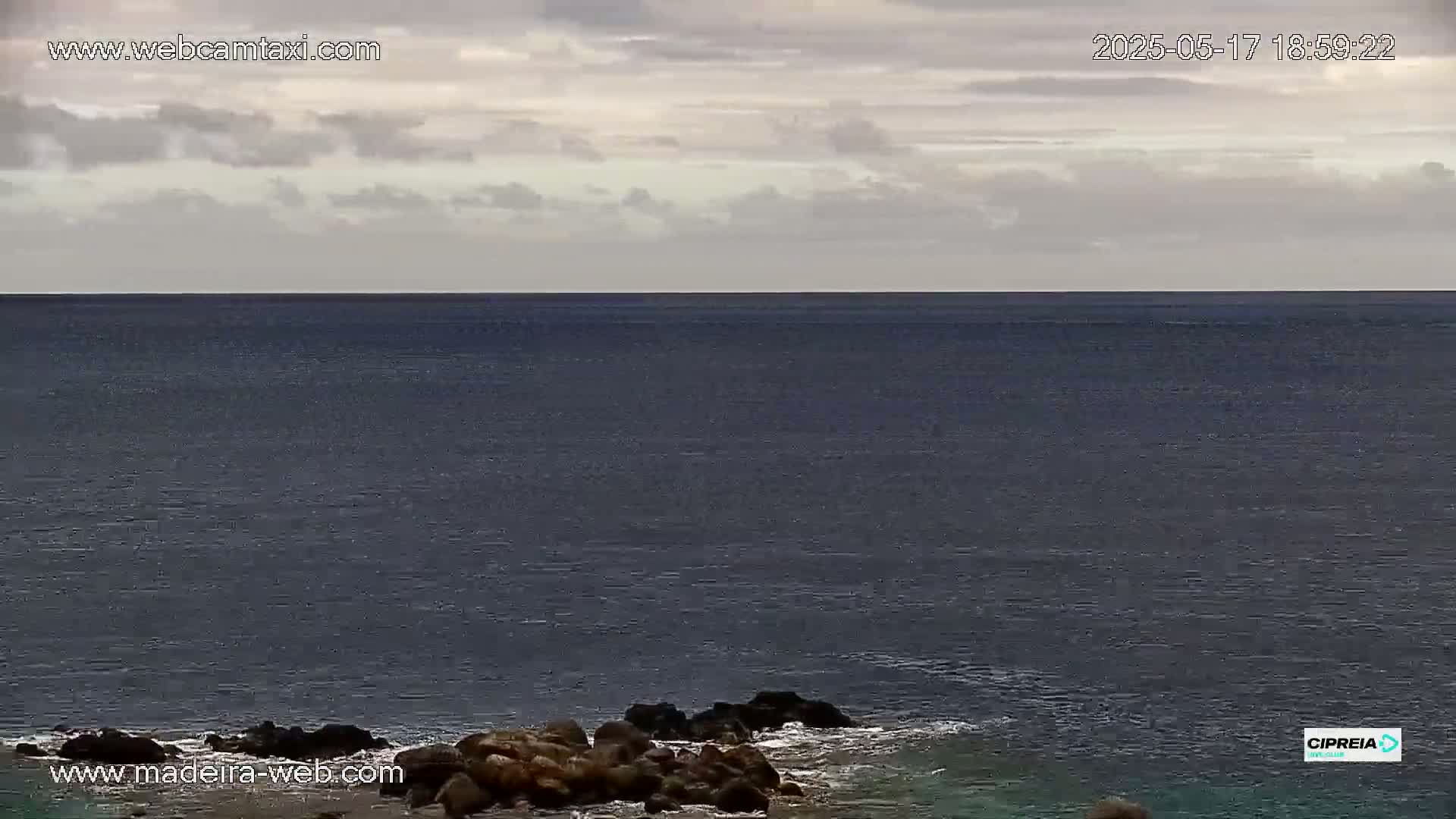 Dark, calm ocean water meets a cloudy sky, with dark rocks visible in the foreground.