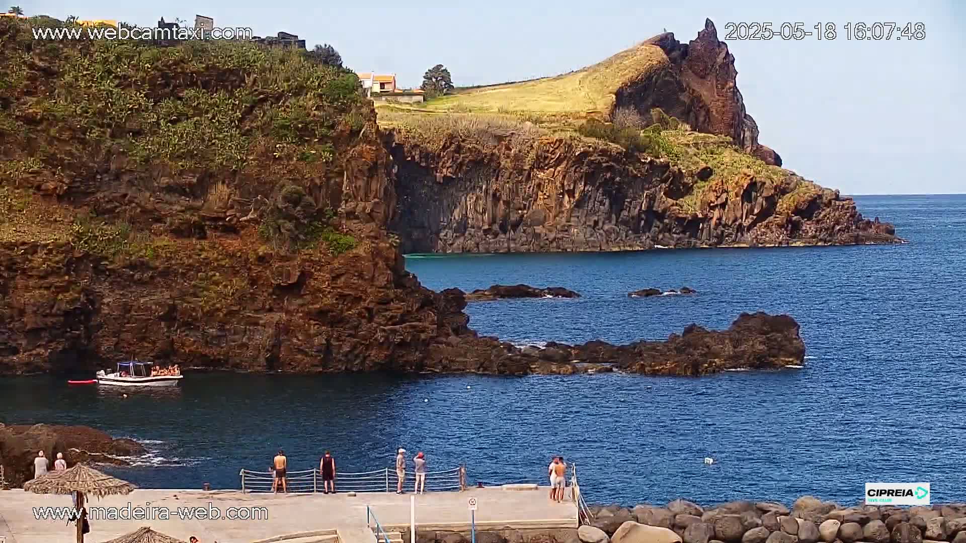 A rocky coastline with dark volcanic cliffs and a calm, blue ocean under a sunny sky, features a small boat and several people standing on a dock.