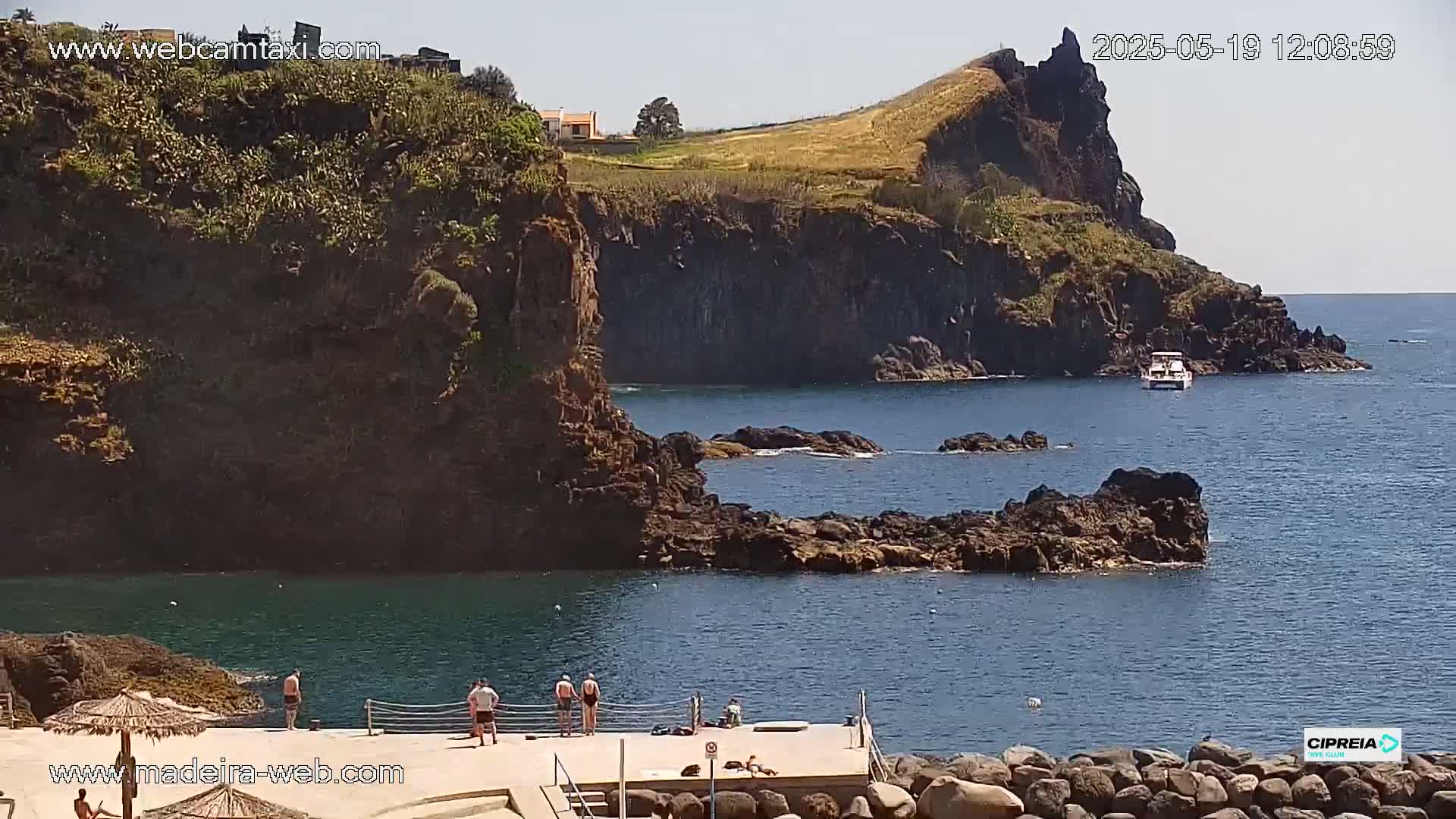 A sunny day reveals a rocky coastline with several people standing on a small dock, a boat offshore, and a small house visible on a distant hill.