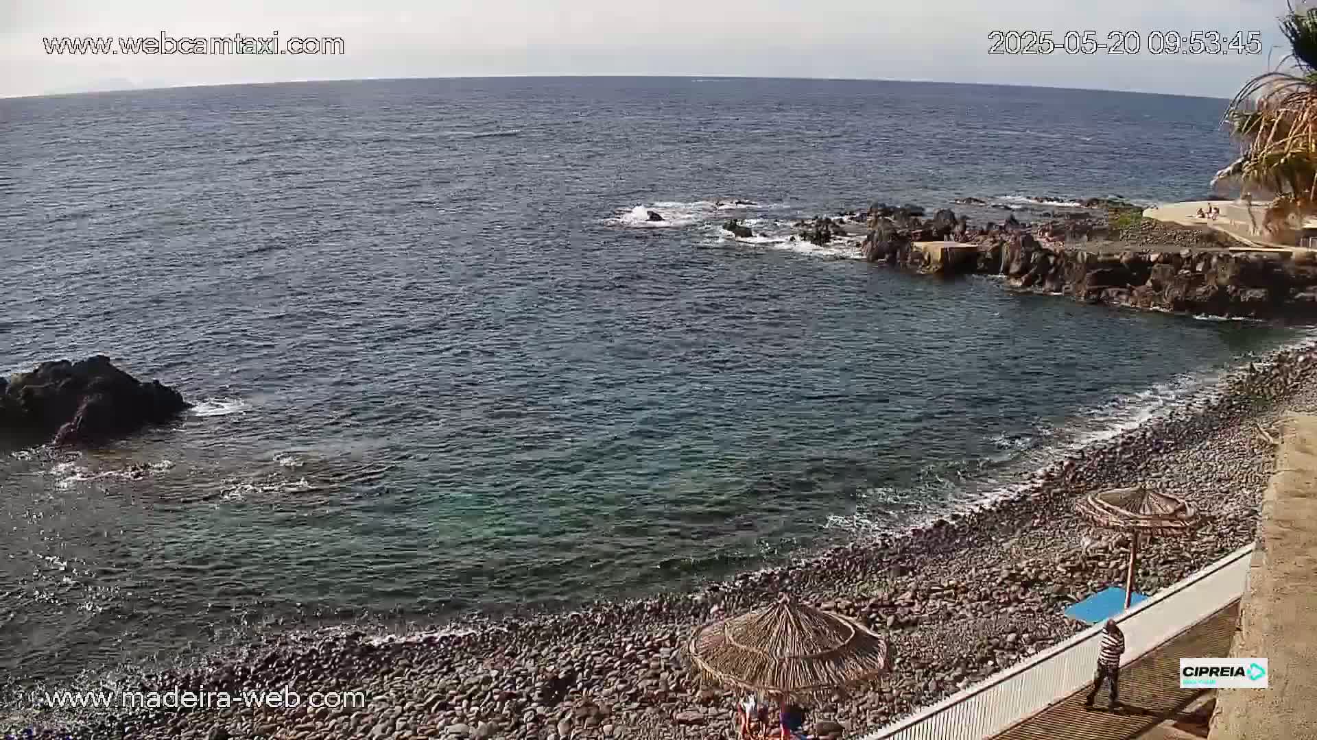 A rocky, pebble beach meets a calm, dark-blue ocean under a mostly sunny sky.