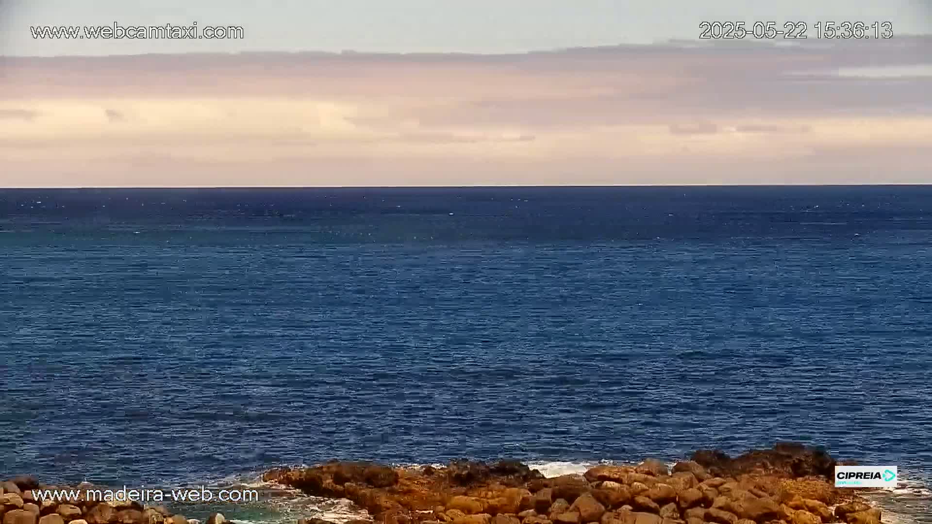 A calm, dark blue ocean meets a pale, cloudy sky at the horizon, with dark rocks visible in the foreground.