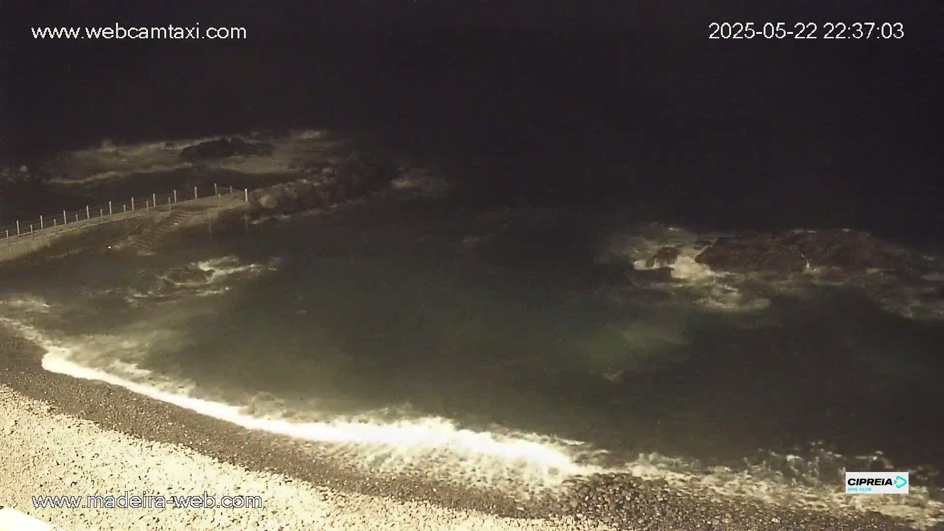 At night, waves crash against a rocky shore and lap at a pebble beach near a dark, calm pool of water enclosed by a fence.