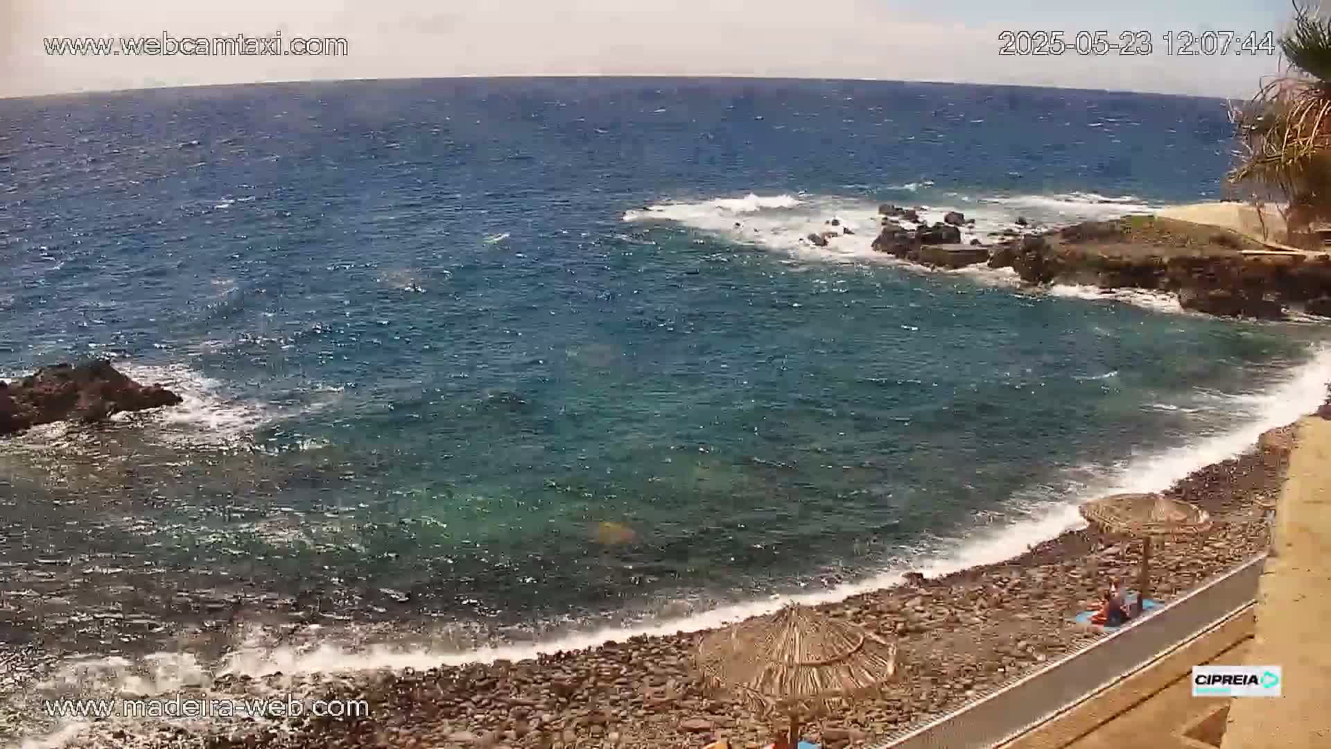 A rocky beach with a few people relaxing under beach umbrellas is lapped by a somewhat choppy sea under a partly cloudy sky.