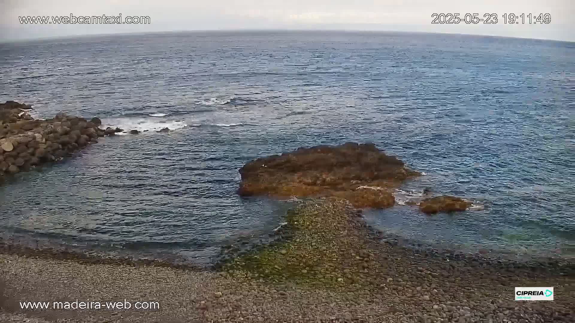 A rocky outcrop in a calm, dark-blue ocean is connected to a pebbled shore by a narrow, submerged rock formation.