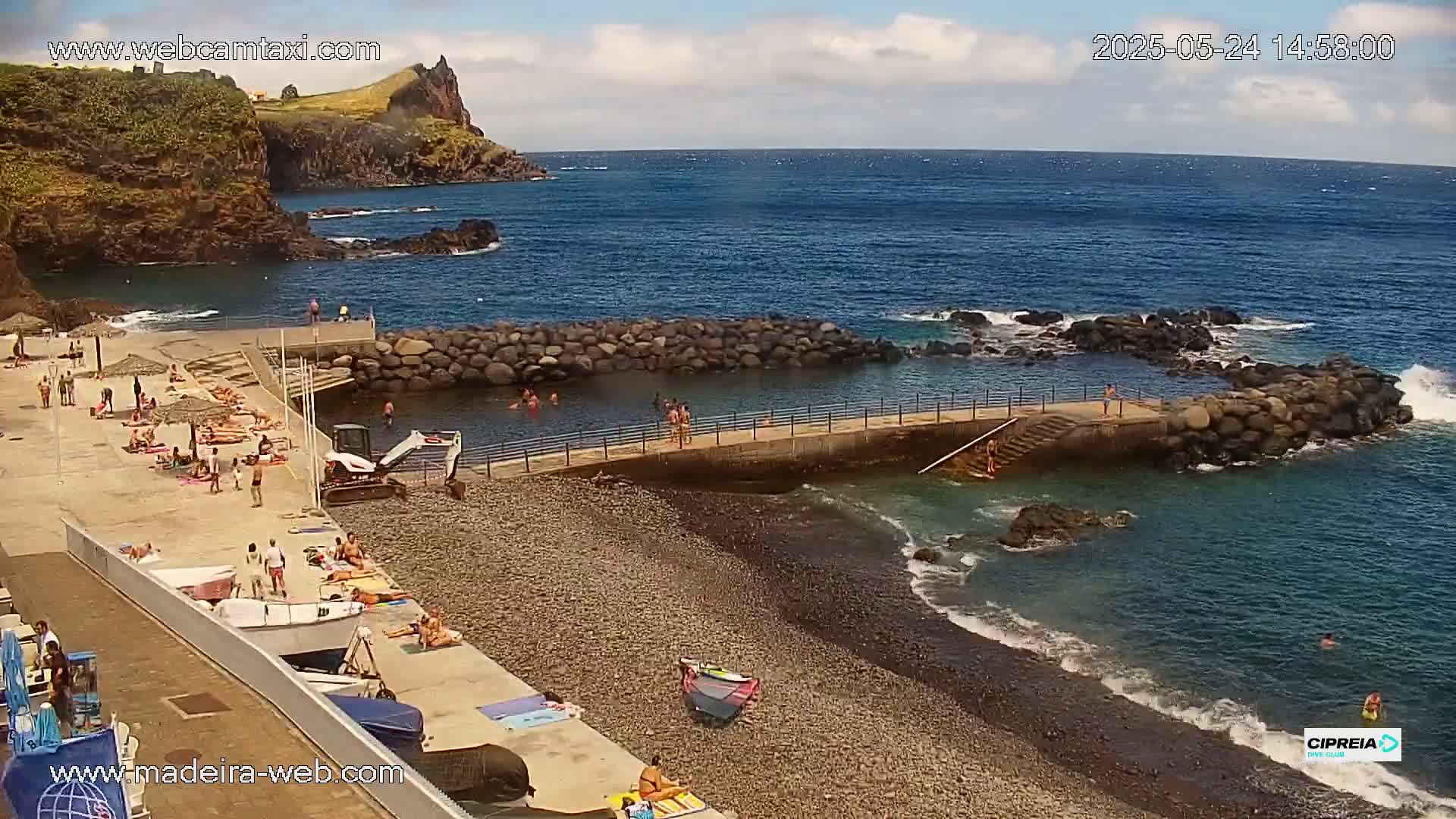 A rocky beach with a calm ocean, a small protected swimming area, and many people sunbathing and swimming on a partly sunny day.