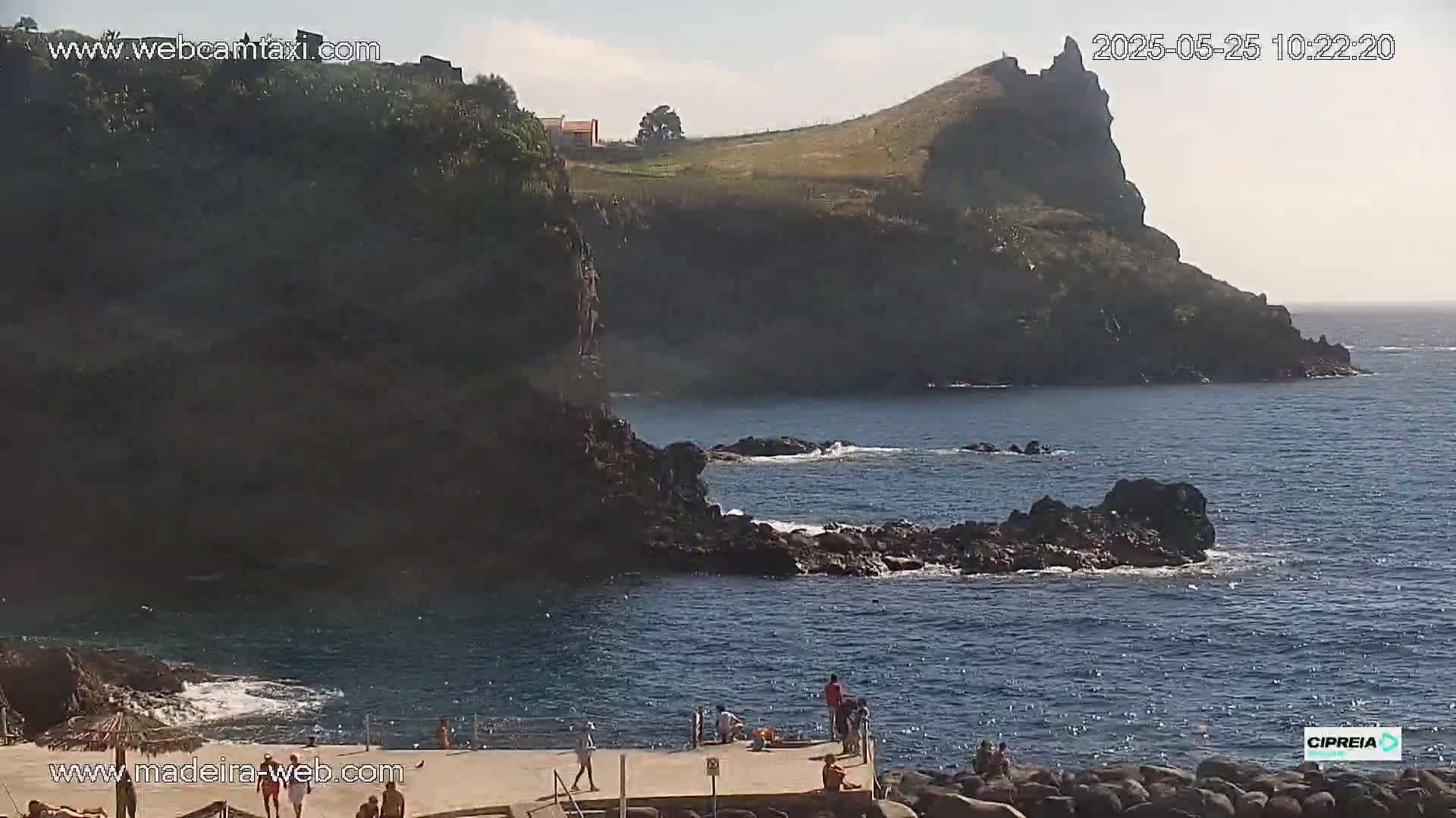 A rocky coastline with a small group of people on a platform overlooking a dark blue ocean under sunny skies.