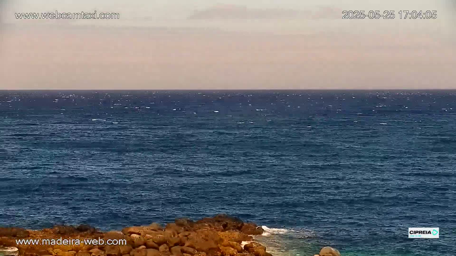 A mostly calm, dark blue ocean meets a rocky shoreline under a light, hazy sky.