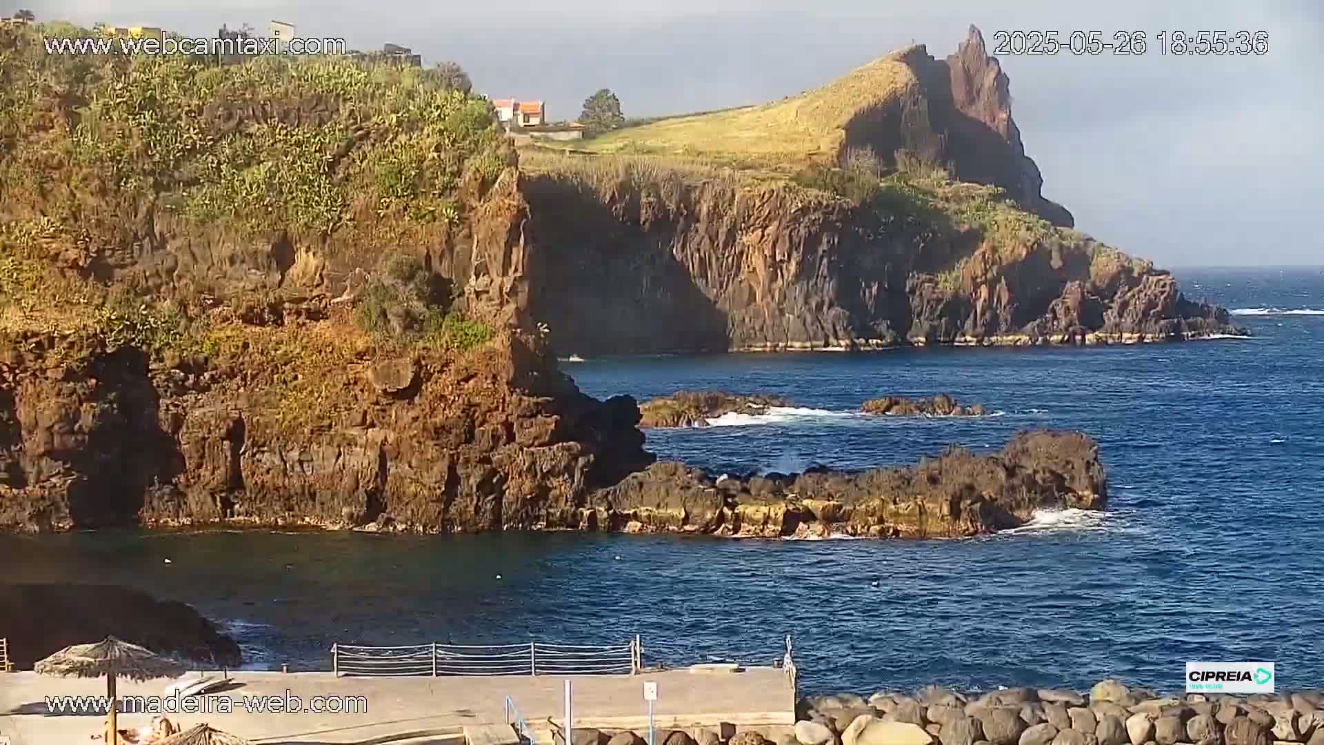 A rocky, cliff-lined coast meets a calm, blue ocean under a mostly clear sky.
