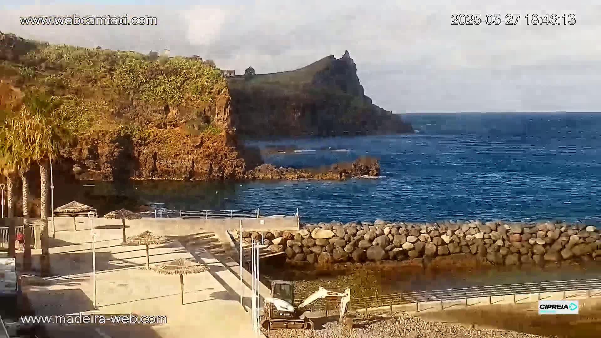 A small, rocky cove with calm ocean water is partially enclosed by a low stone wall, and a small construction vehicle is visible near a paved area with a few beach umbrellas.