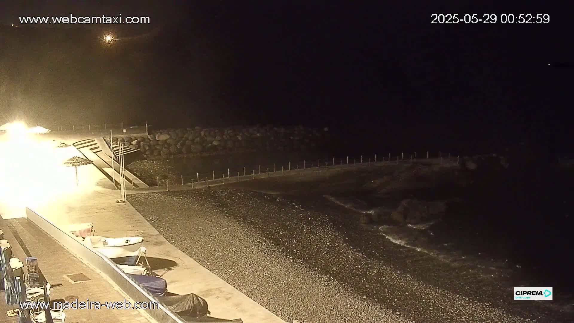 A dark, nighttime beach scene shows a pebble shoreline, calm water, and small boats docked near a lit walkway.