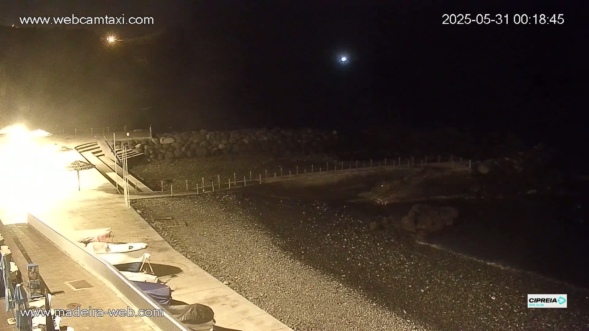 A nighttime view of a pebble beach with boats and a walkway under a mostly clear sky.