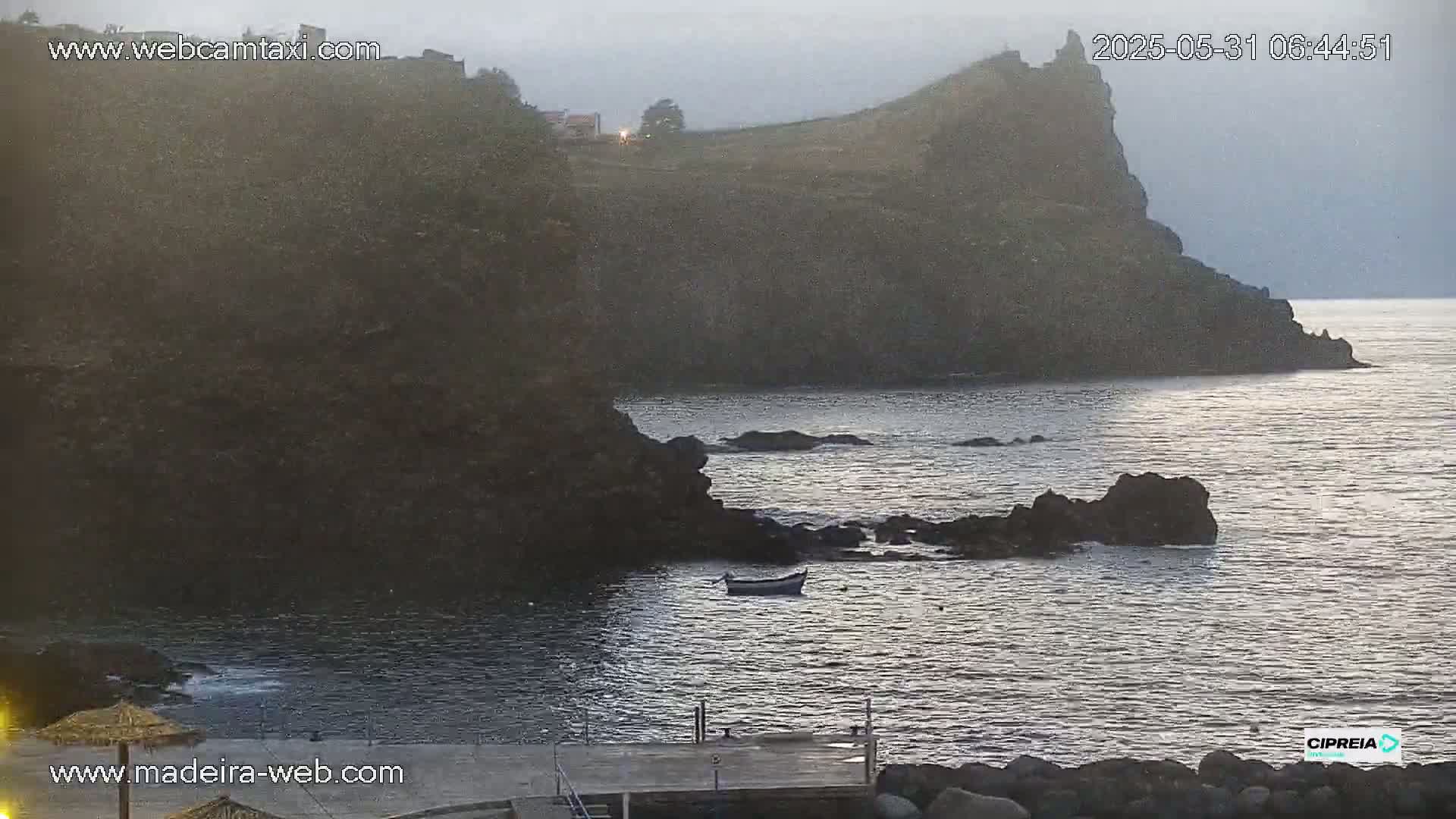 A small boat rests in calm, dark water near a rocky, dark coastline under an overcast sky.