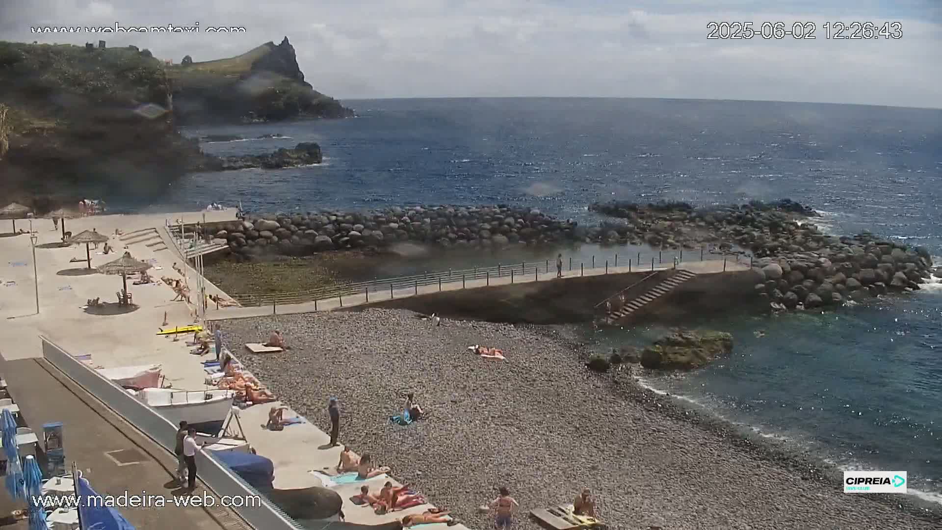 A pebble beach with people sunbathing and swimming in a calm, partly sunny ocean next to a rocky cliff.