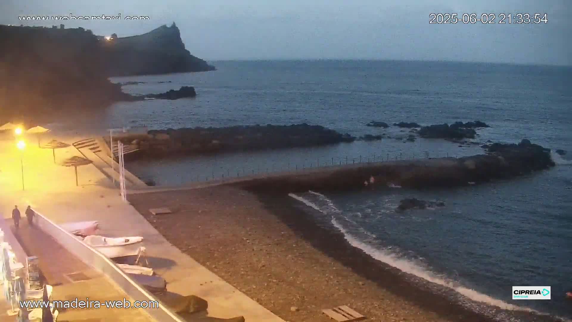 A dark, pebble beach at dusk is partially enclosed by a low stone wall and has a few small boats beached nearby, with a calm ocean and a cliff in the background.