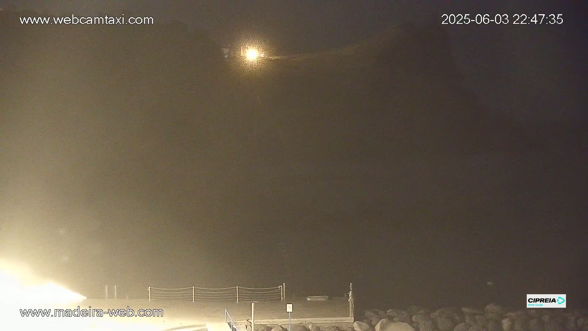 A hazy night scene shows a dark body of water, a low fence, and some rocks at the water's edge, illuminated by a bright light source in the distance.