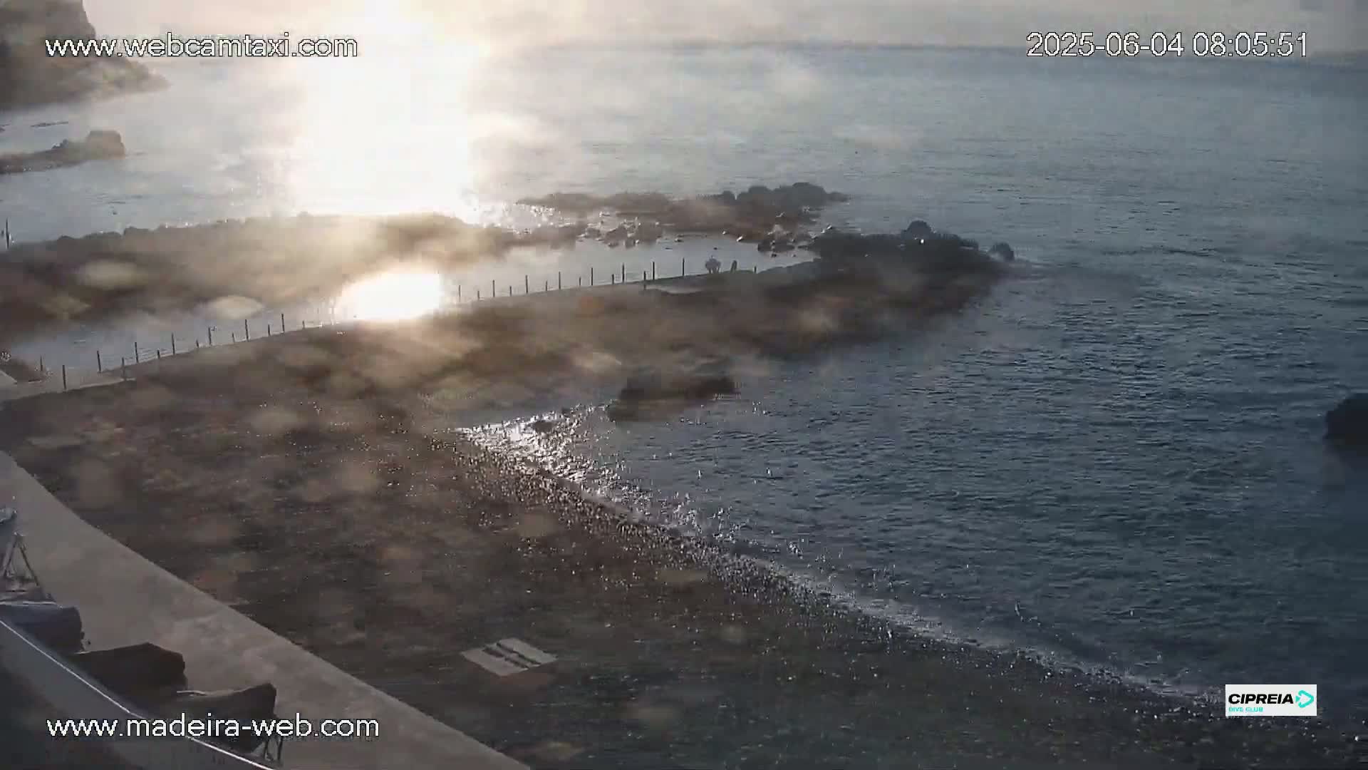 A hazy sunrise illuminates a rocky shoreline with a calm ocean and a small enclosed pool.