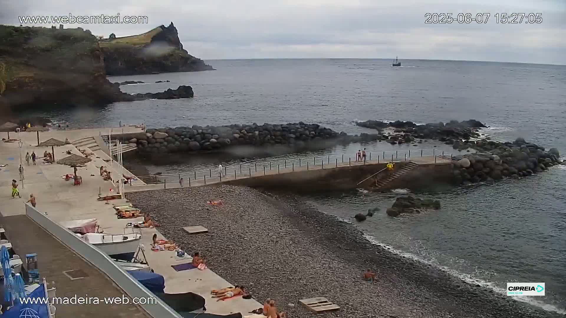 A rocky beach with a concrete walkway and several people sunbathing and swimming, under an overcast sky.