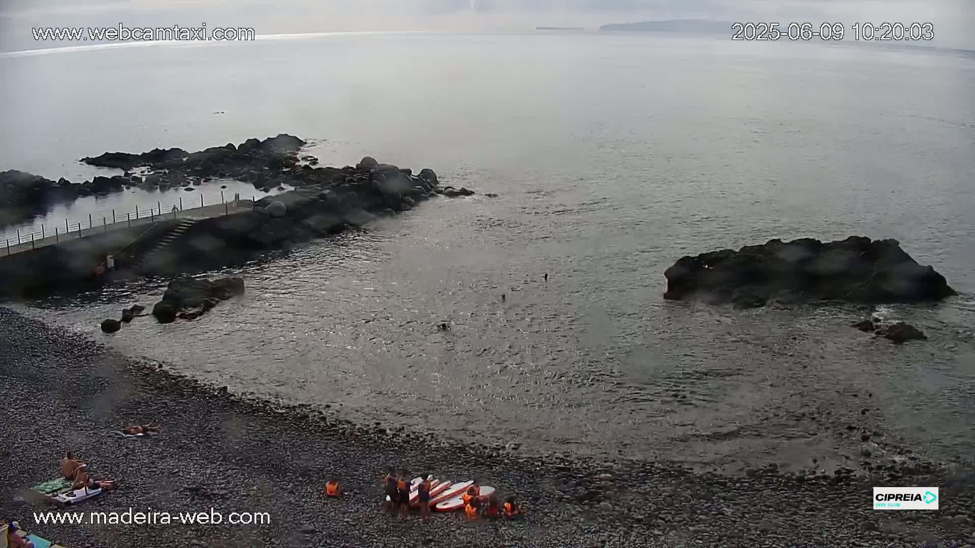 A rocky, pebble beach with a small group of people near paddleboards in calm, overcast water.