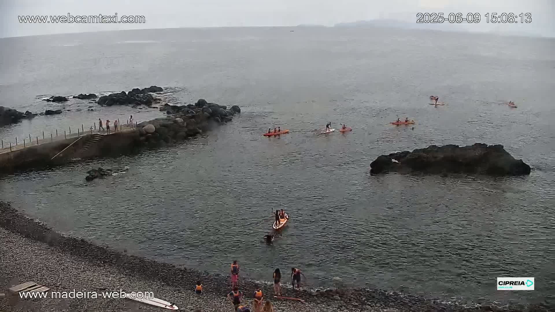 A rocky coastline with several people kayaking and paddleboarding in calm, overcast waters.