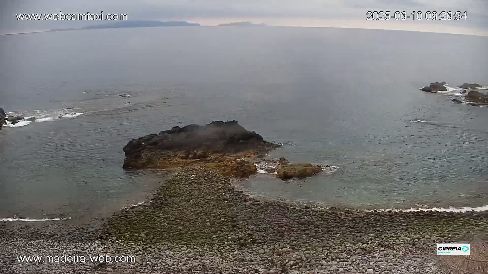 A rocky shoreline with dark, jagged rocks extends into a calm, gray-green ocean under an overcast sky.