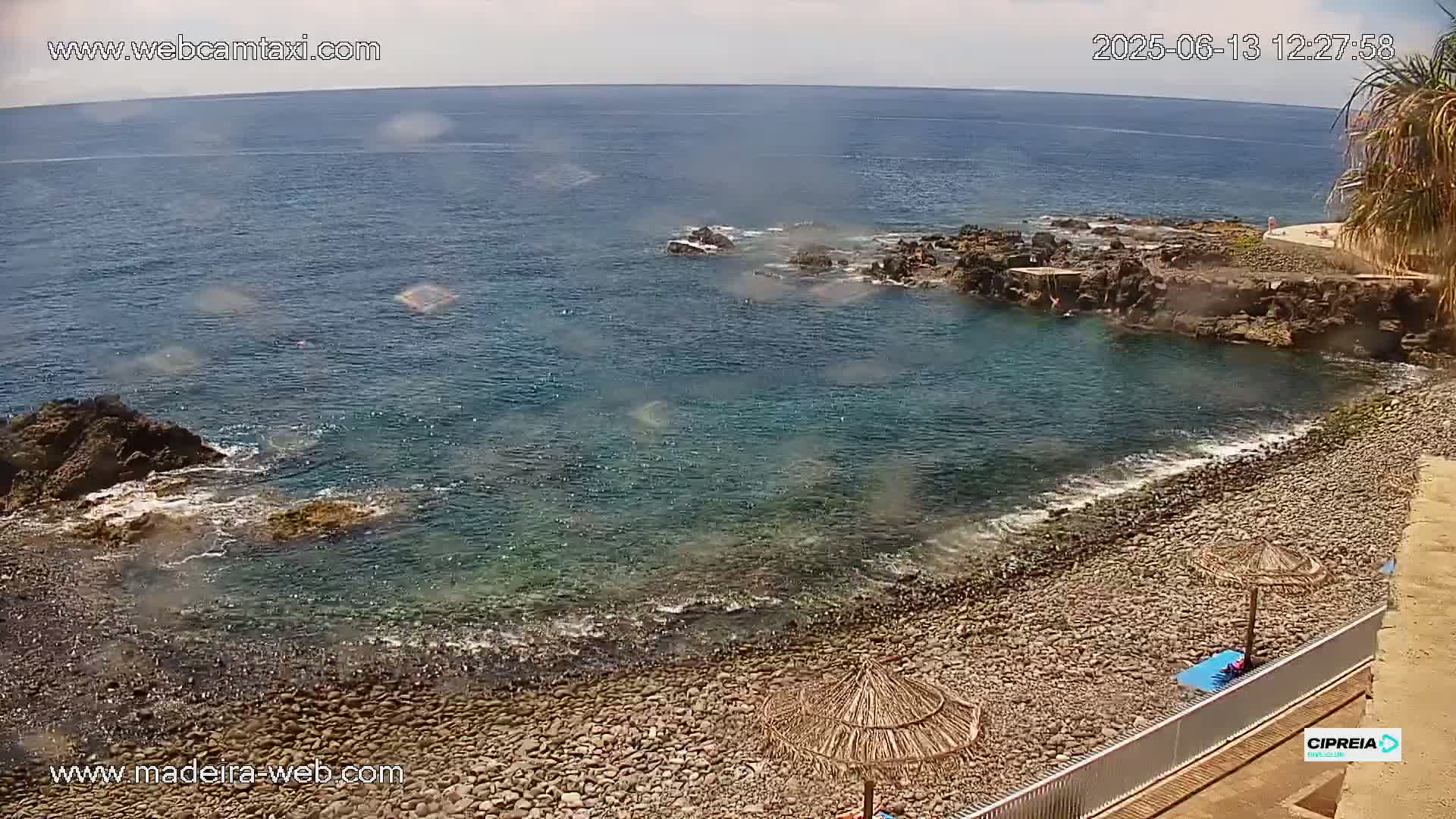 A rocky beach with calm, clear water laps gently against the shore under a sunny sky.