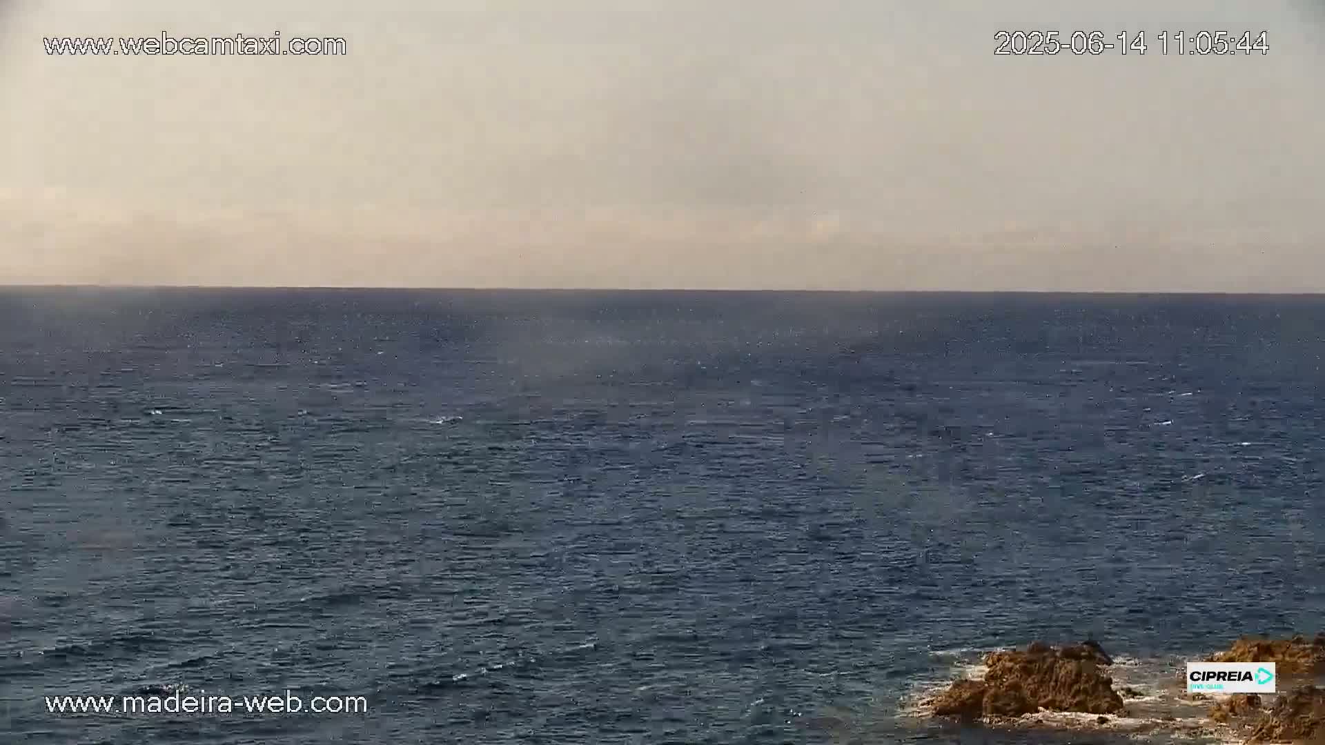 A slightly hazy, calm ocean stretches to the horizon, with dark rocks visible in the foreground.
