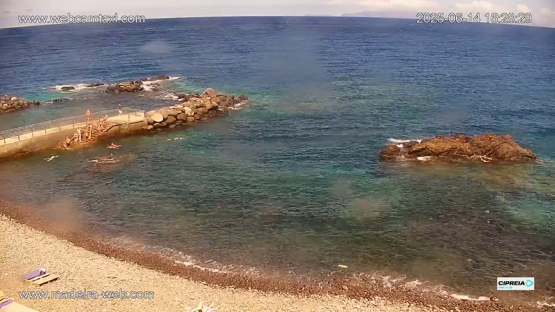 A rocky beach with calm, clear turquoise water, several people swimming near a small stone jetty under a mostly sunny sky.