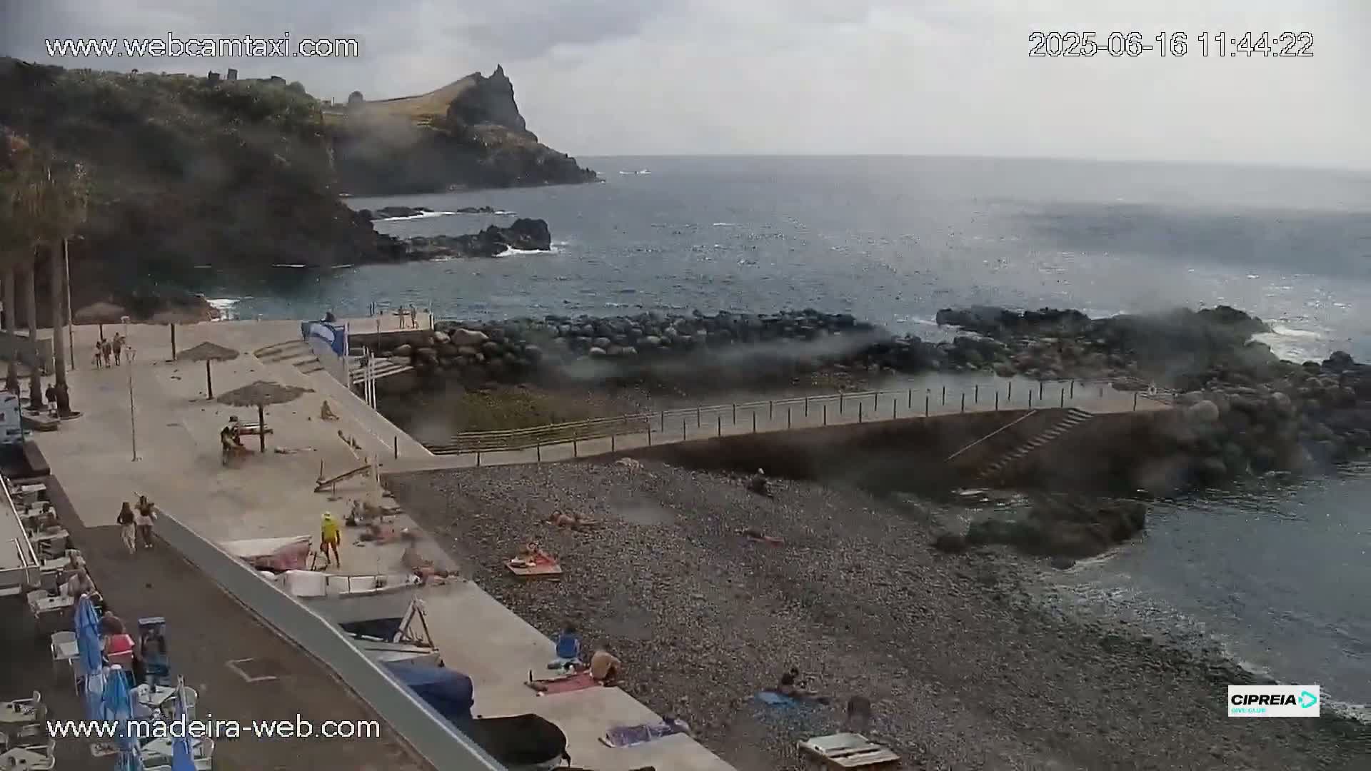 A rocky beach with people sunbathing and swimming, a small harbor, and a dark, overcast sky over a choppy ocean.