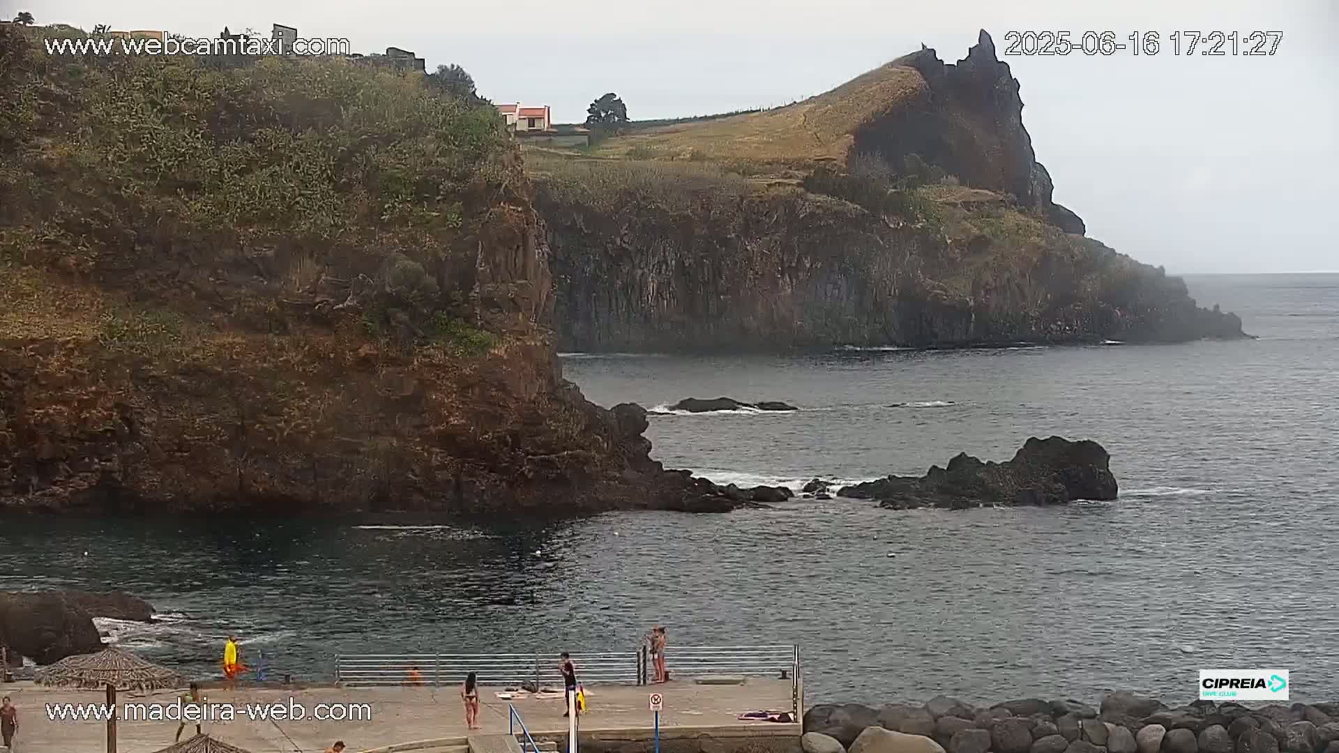 A rocky coastline with a small, calm cove where several people are swimming and lounging on a dock under an overcast sky.
