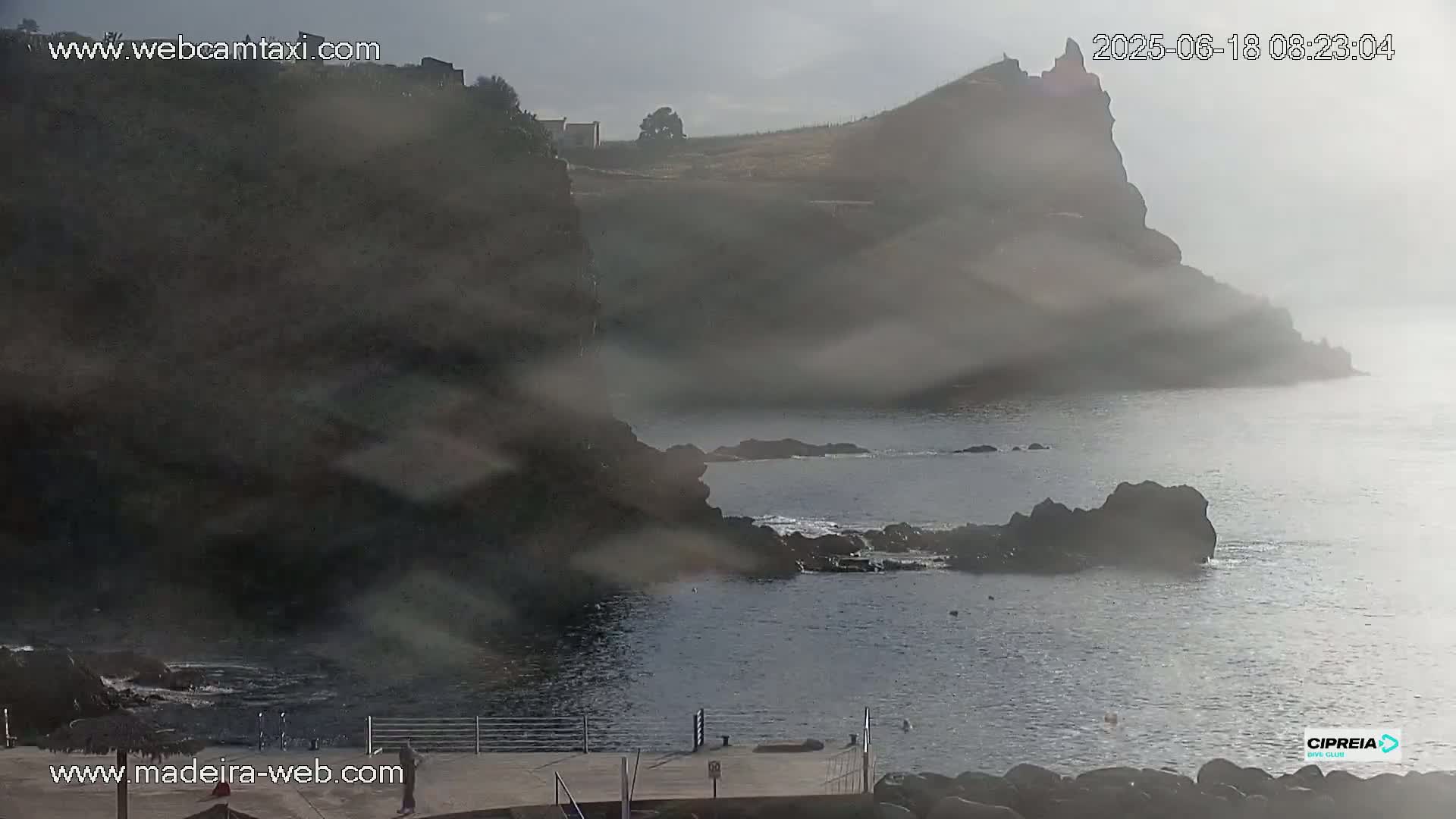 A hazy, overcast day reveals a rocky coastline with a small structure atop a cliff overlooking a calm body of water.