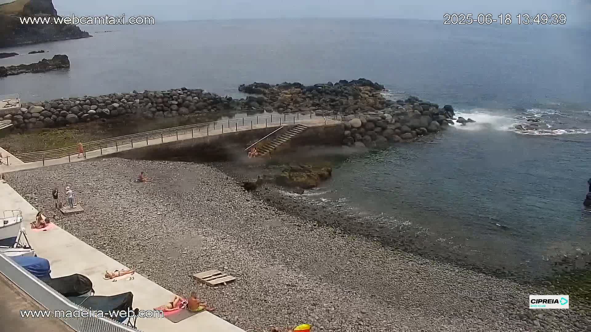 A rocky beach with calm water and several people relaxing and swimming, under an overcast sky.