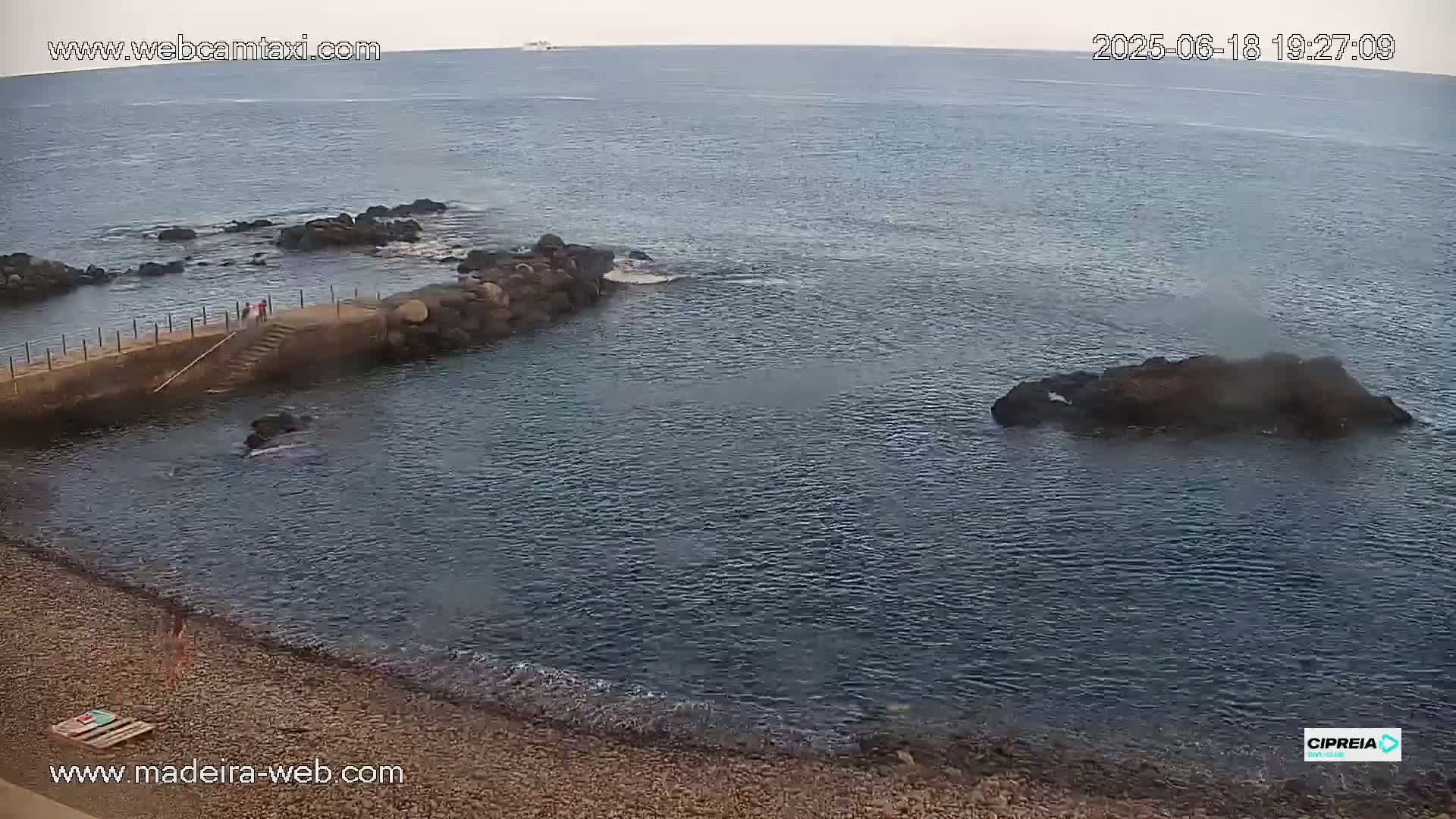 A rocky shoreline with a small pebble beach and calm, dark-blue water under an overcast sky.