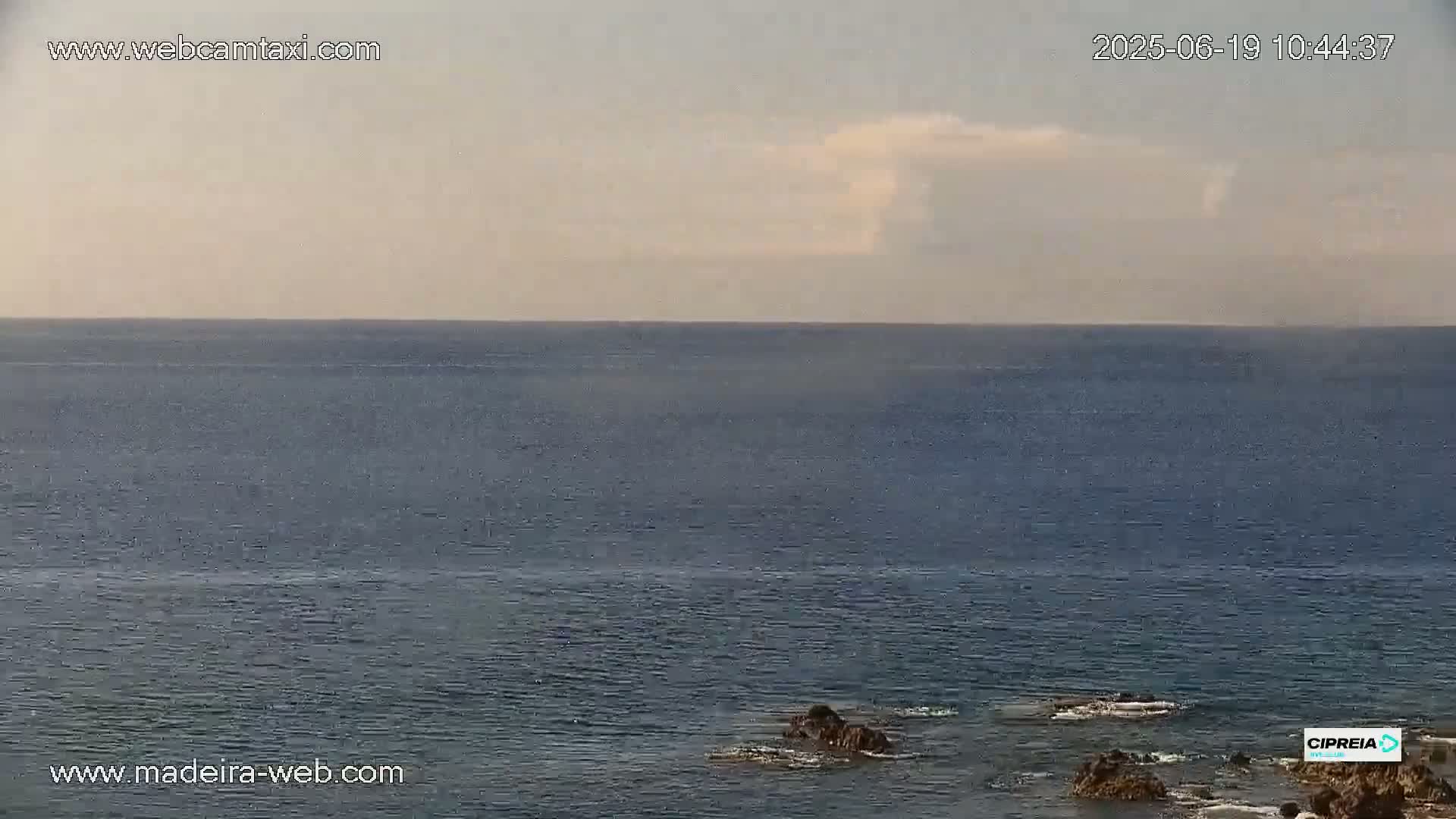 A calm ocean with a few dark rocks visible in the foreground under a mostly clear, light-grey sky.