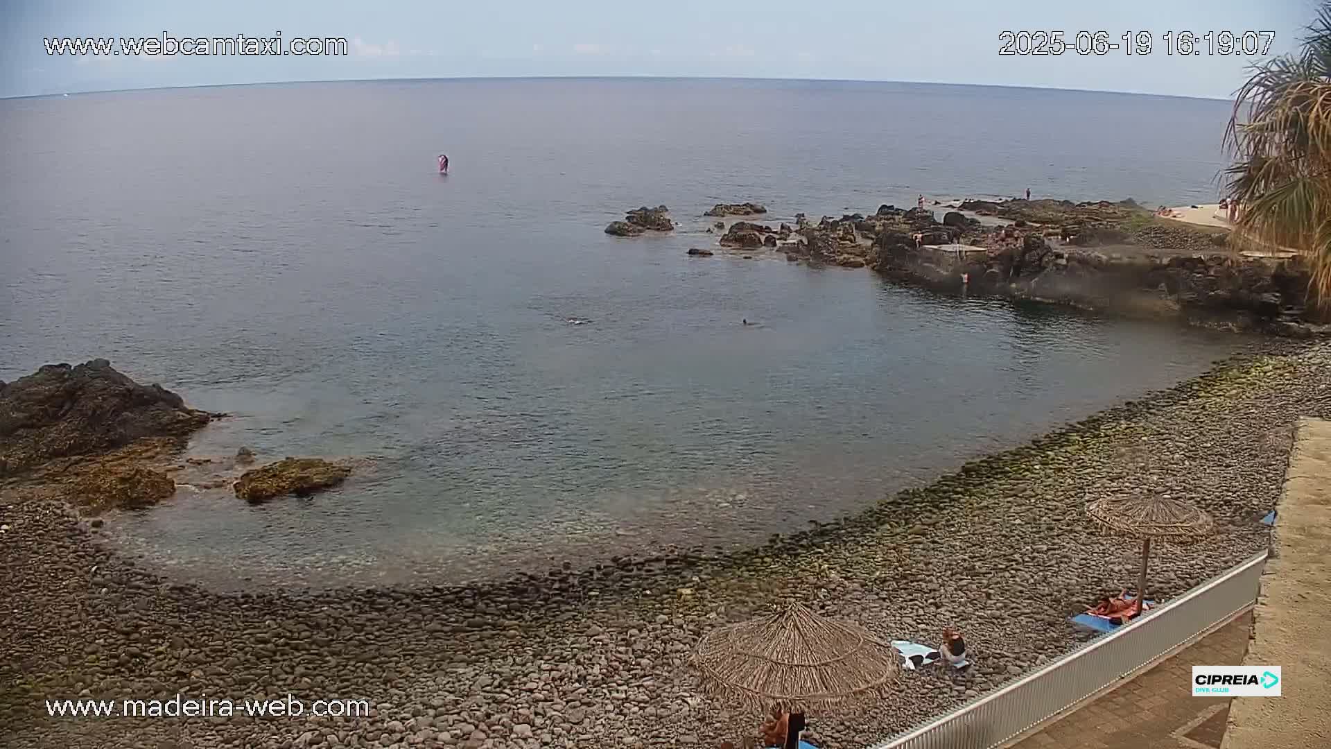 A rocky, pebble beach with calm, clear water and a few people swimming and sunbathing under a mostly clear sky.