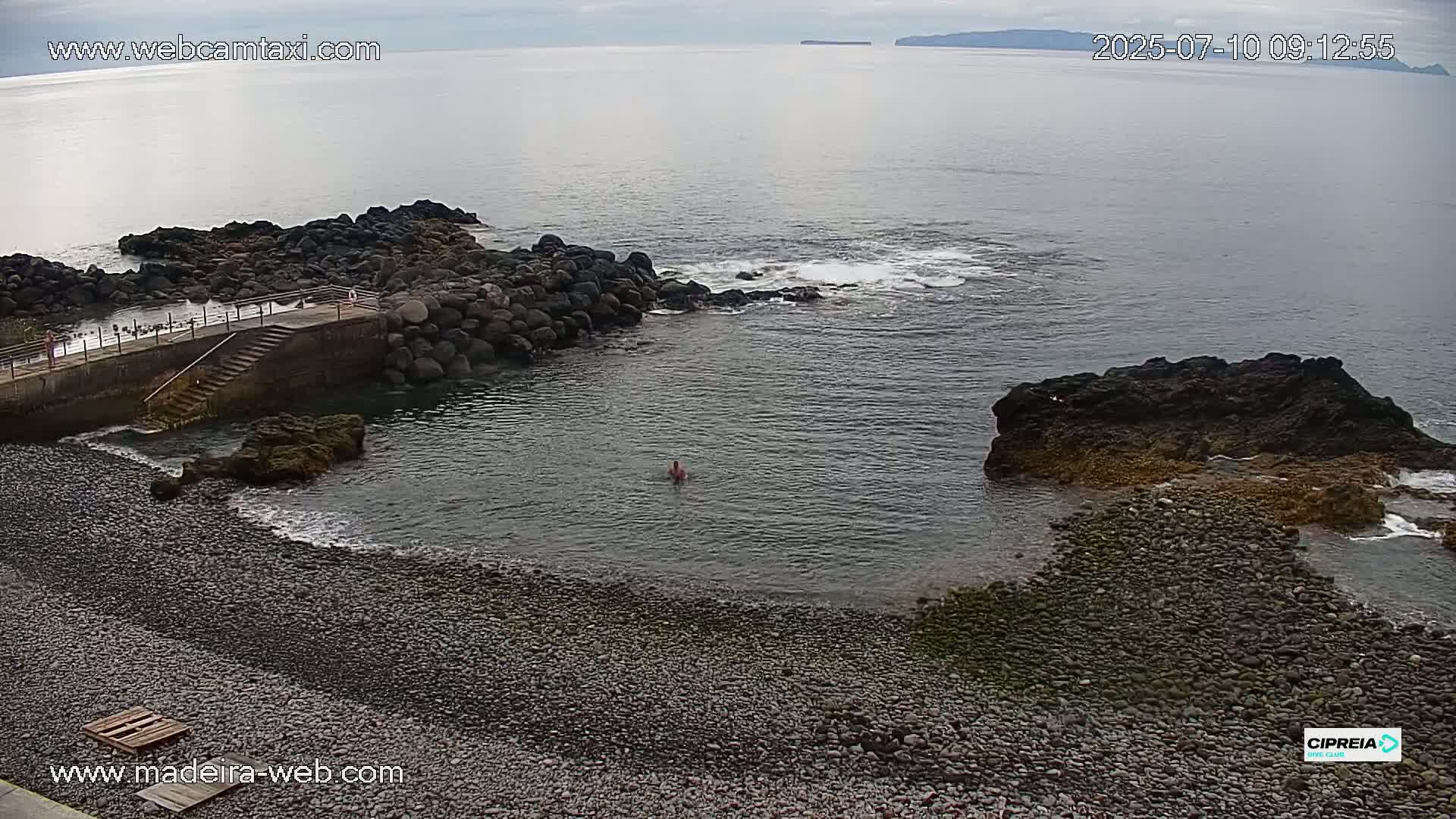 A rocky, pebble beach with a small, calm cove features a person swimming and a concrete staircase leading to a pier under an overcast sky.