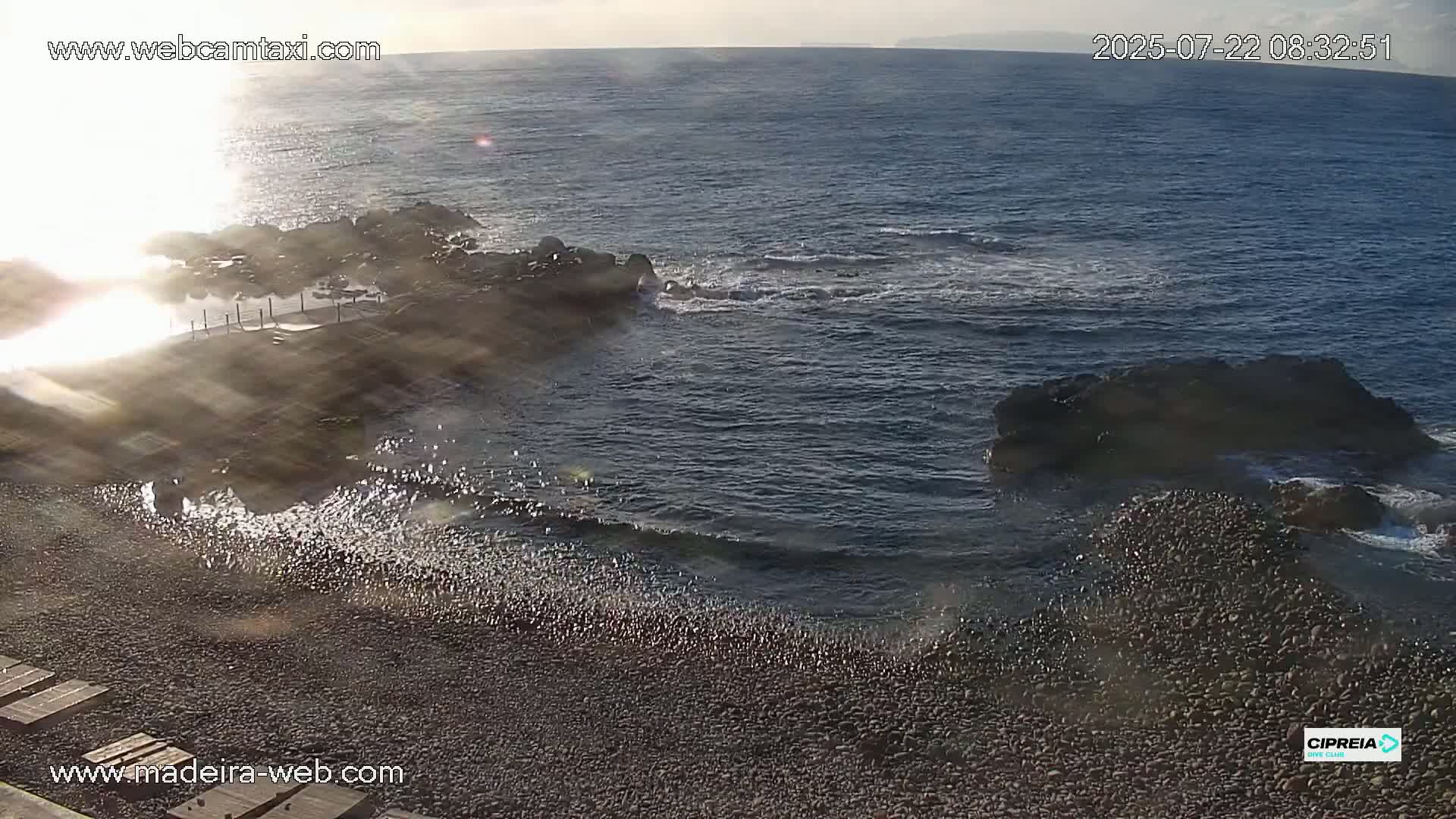 A rocky shoreline with a pebble beach meets a calm ocean under a partly sunny sky.