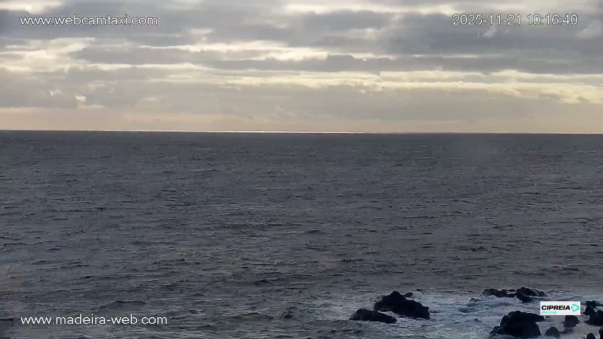 A choppy, dark ocean extends to a bright horizon under a partly cloudy sky, with waves breaking on dark rocks in the foreground.
