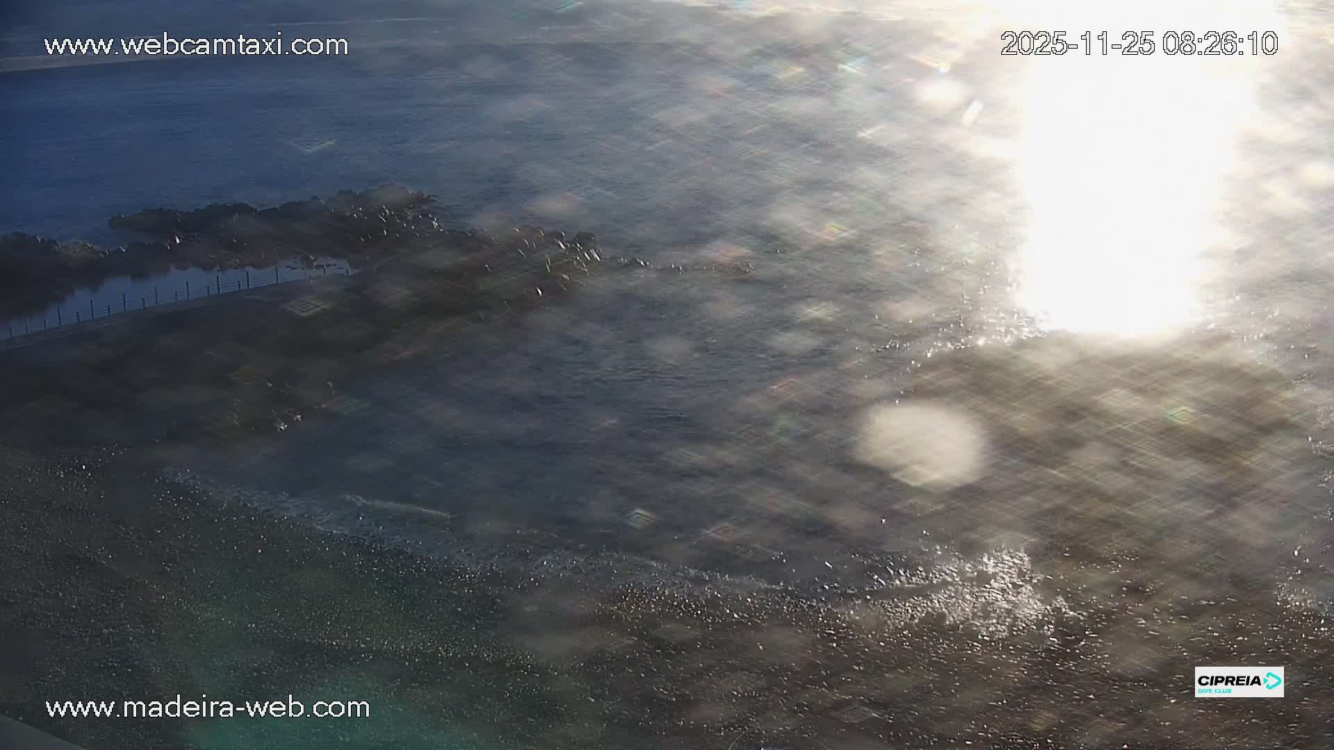 A bright sun glares intensely over a dark, choppy sea with a rocky coastline and a paved promenade visible on the left, under clear weather conditions.